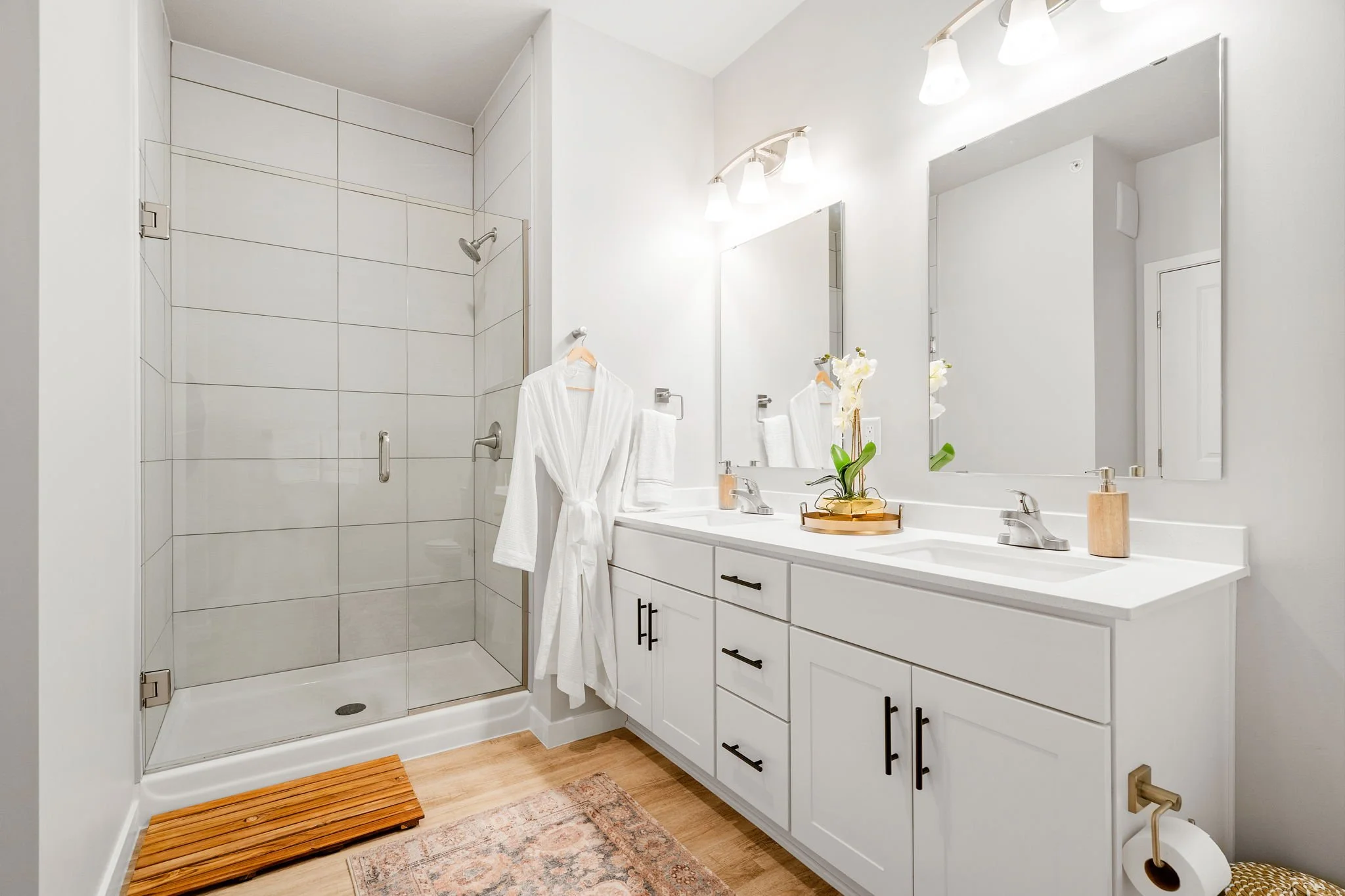 A modern bathroom featuring a glass-enclosed shower on the left, a white double vanity with black hardware in the center, and large mirrors above each sink. Decor includes a tray with a potted orchid, a soap dispenser, and a bathrobe hanging on a hoo