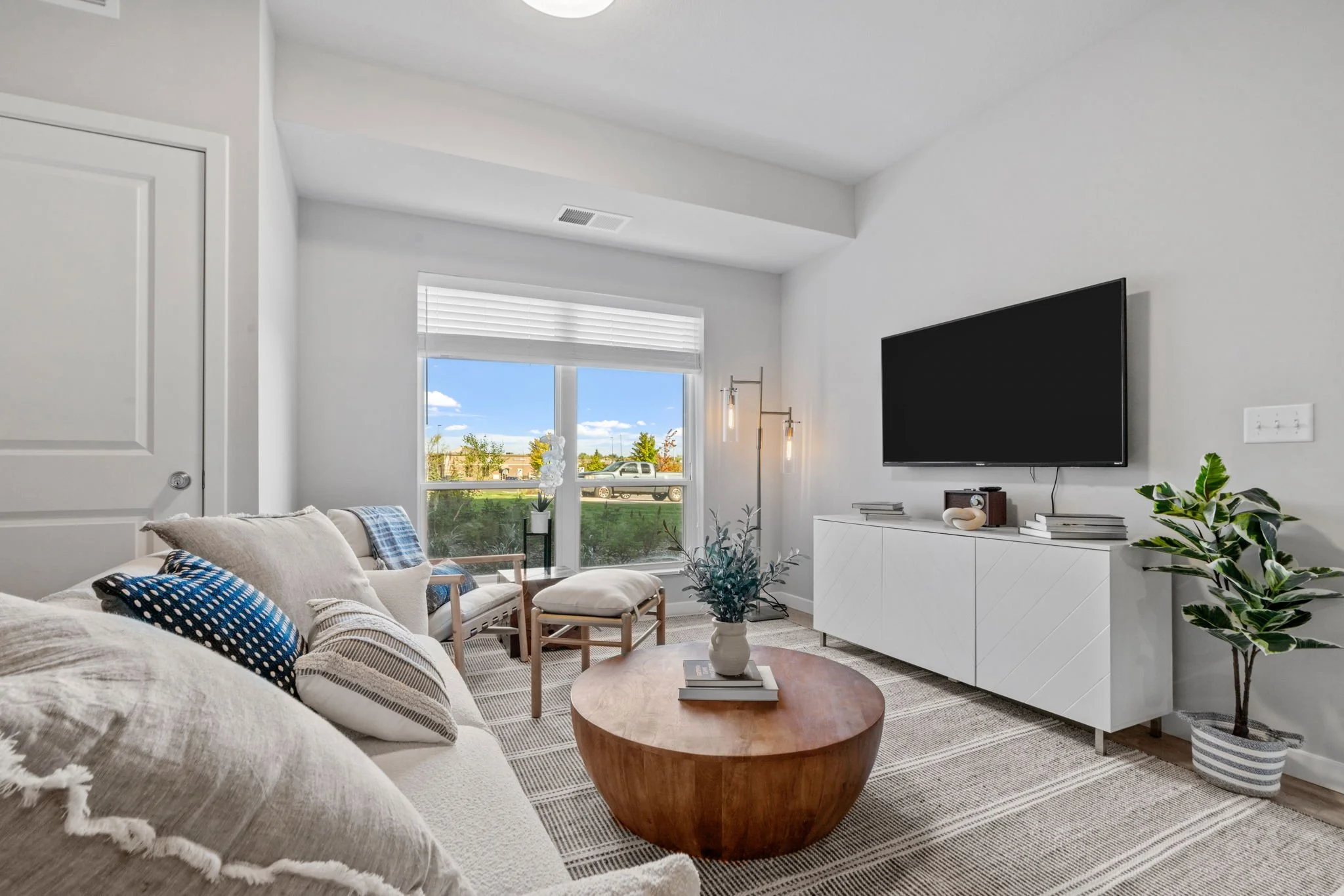 Bright living room with white walls, a beige sofa with patterned pillows, a round wooden coffee table with a vase and books, a large window with a view of the outdoors, and a white entertainment console with a mounted flat-screen TV.