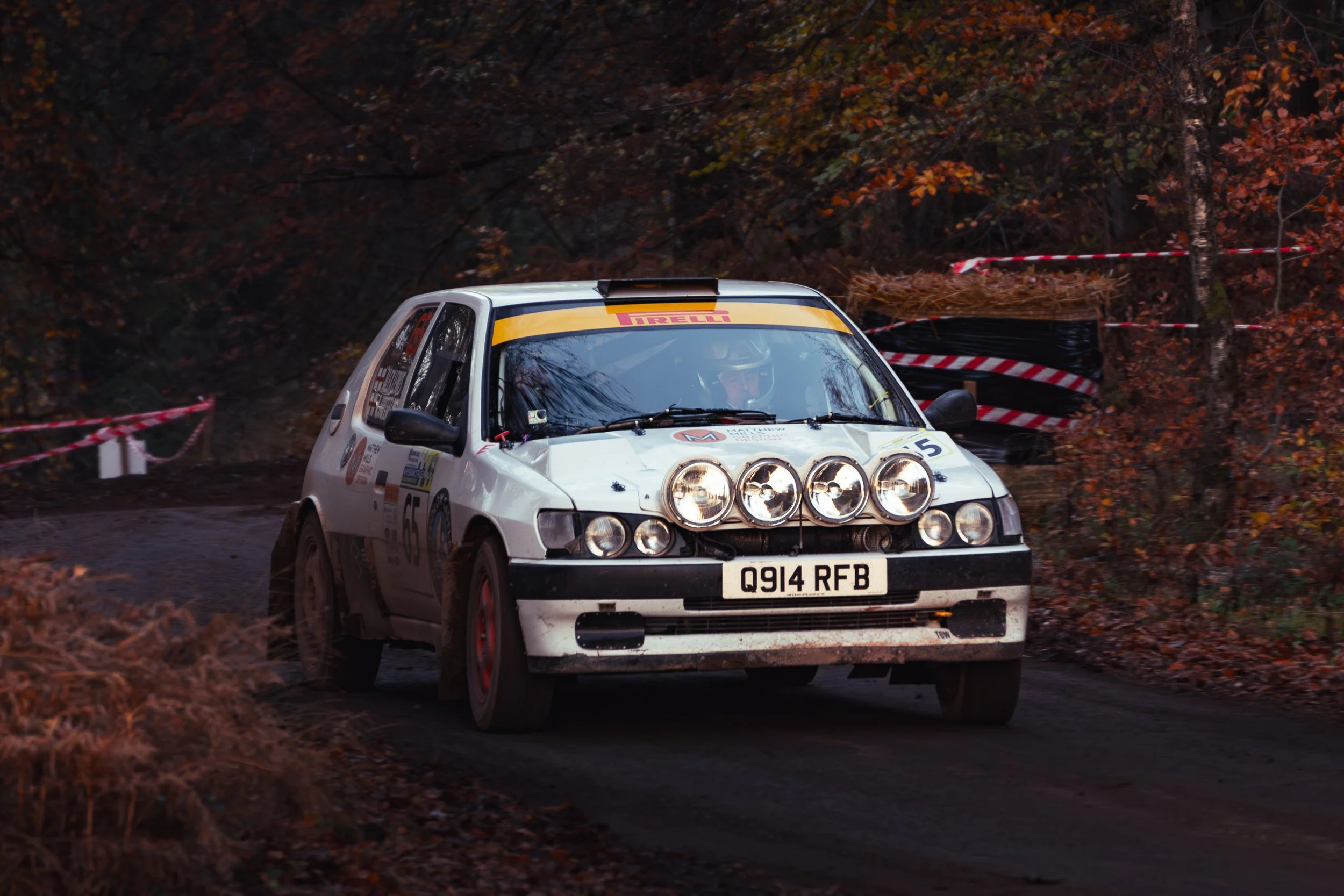 A white rally car with four large front-mounted spotlights and the number 65 on its side, racing on a dirt road through a wooded area with autumn-colored foliage. wye dean rally 