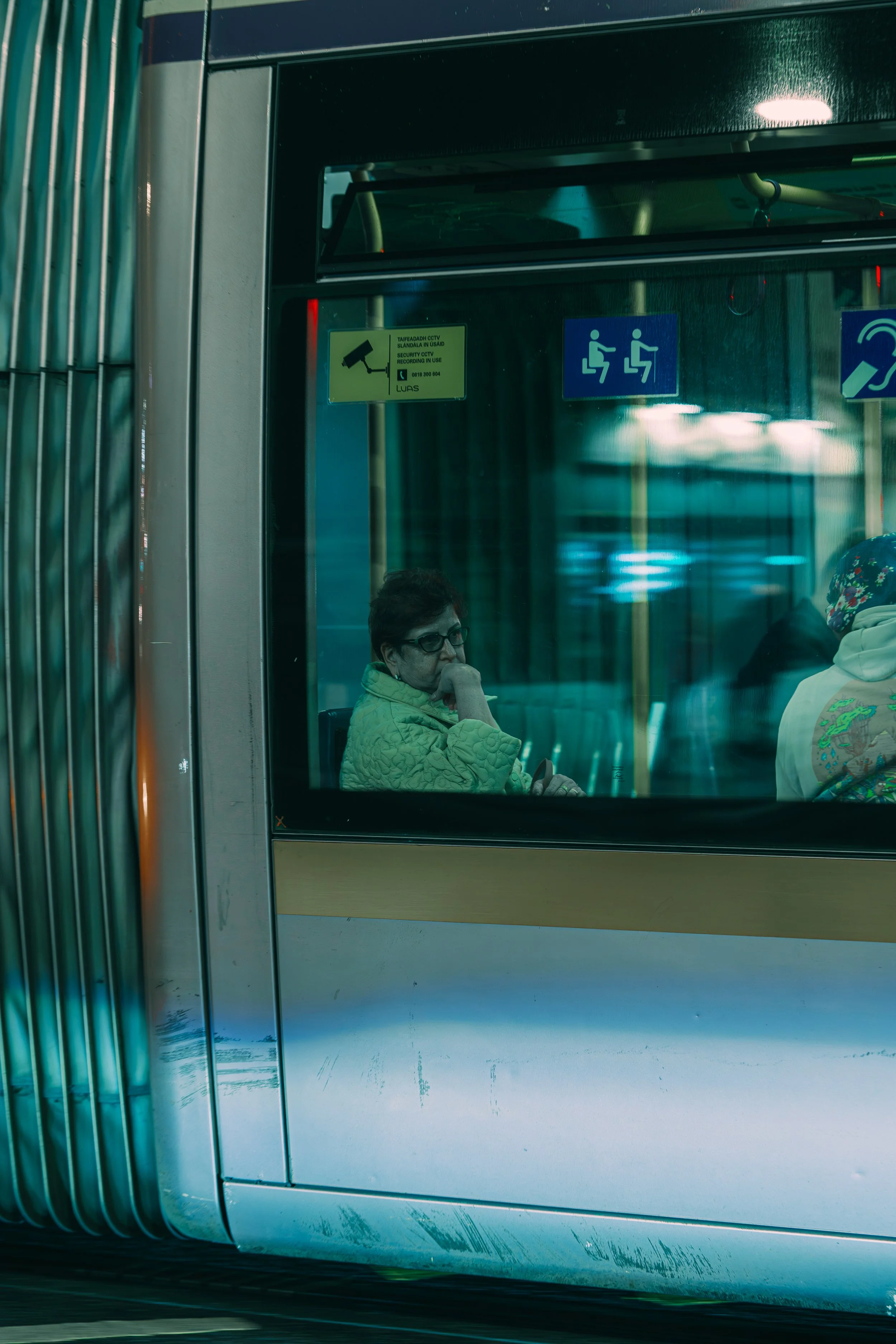 A woman sitting inside a bus, seen through the window, wearing glasses and a yellow quilted jacket.