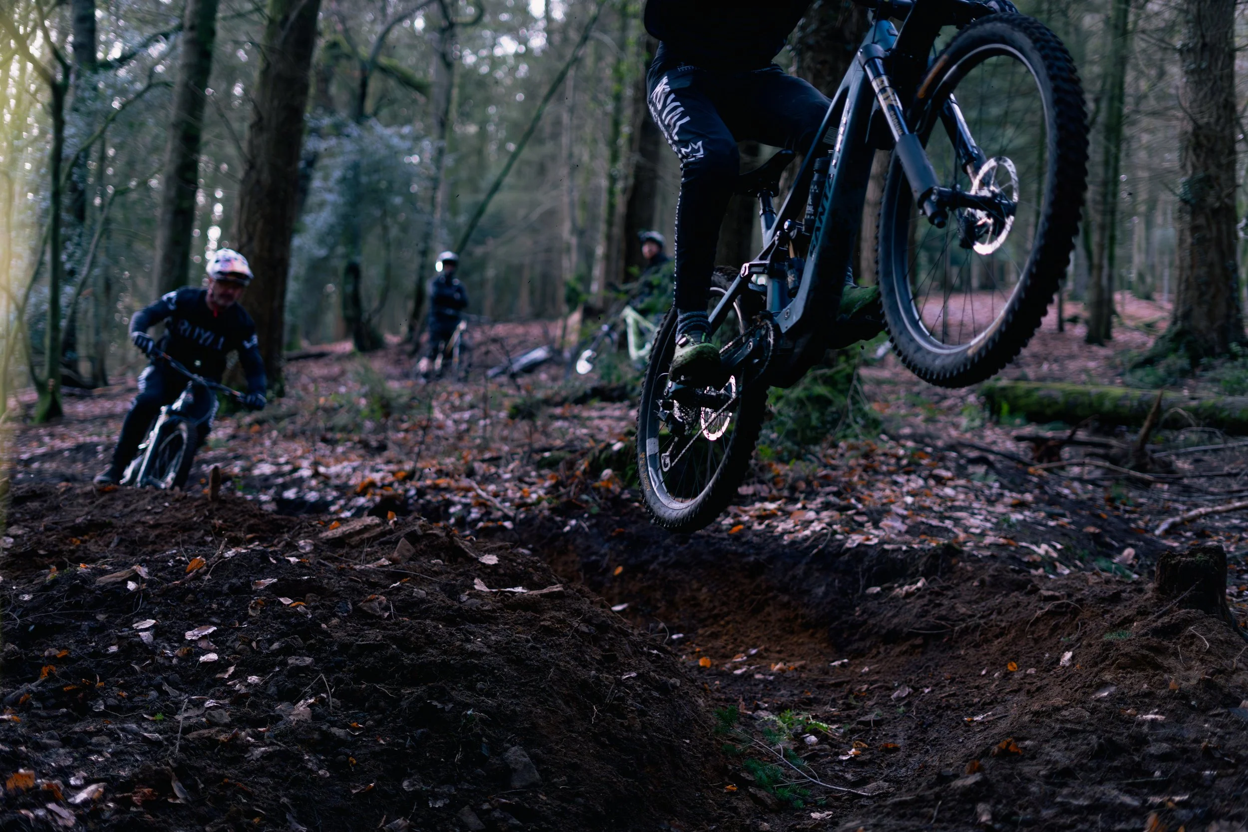 Three mountain bikers in black attire and helmets riding through a wooded forest trail, with one rider airborne over a dirt mound.