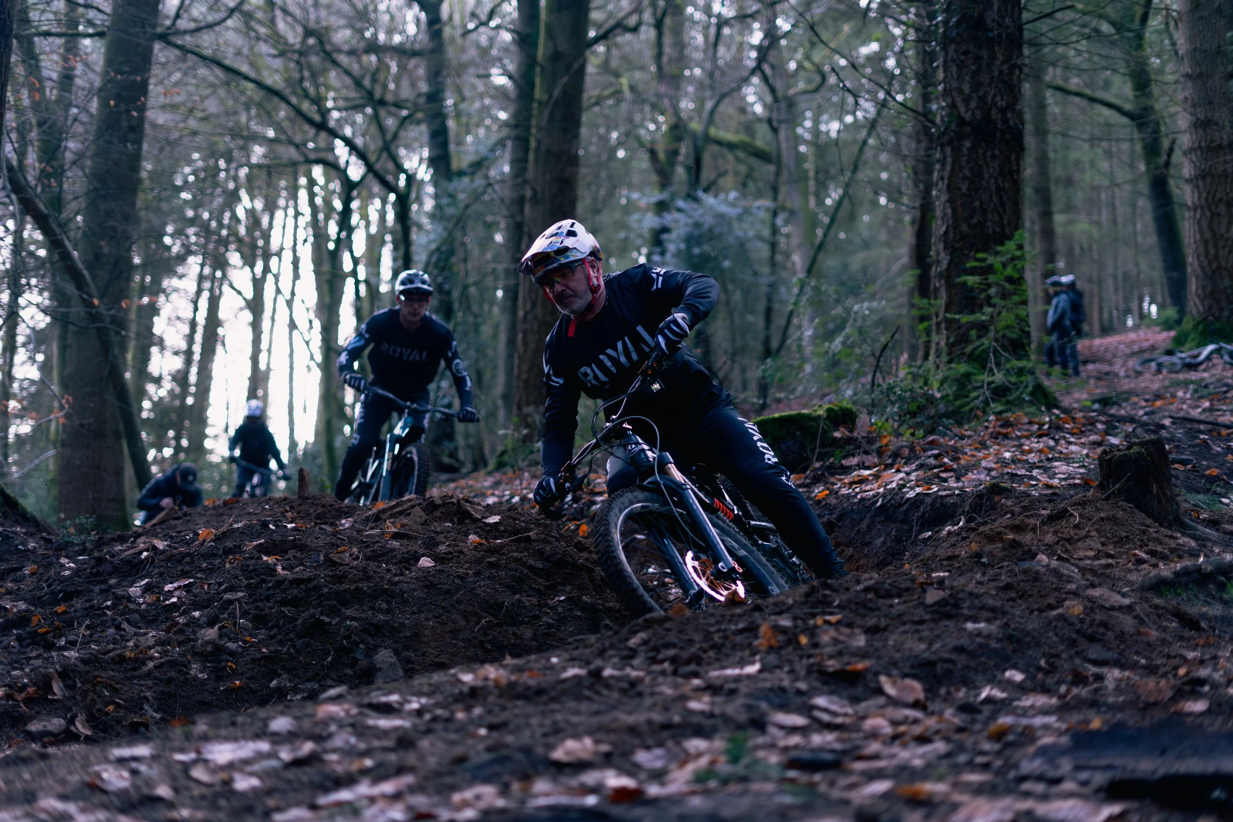 Mountain bikers riding downhill on a dirt trail through a forest with tall trees and fallen leaves.