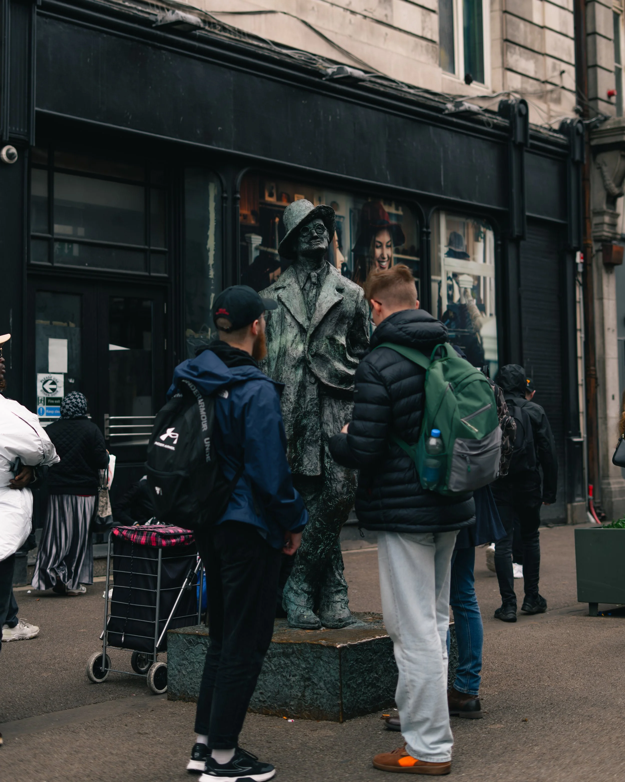 People gathered around a bronze statue of a man in a coat and hat on a city street.