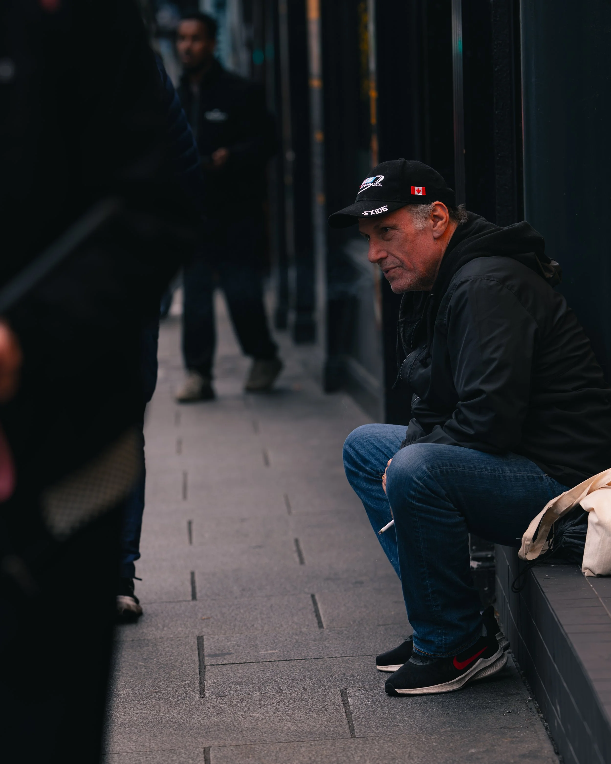 A middle-aged man with gray hair sits alone on a bench on a city sidewalk, wearing a black jacket, jeans, and sneakers, looking contemplative.