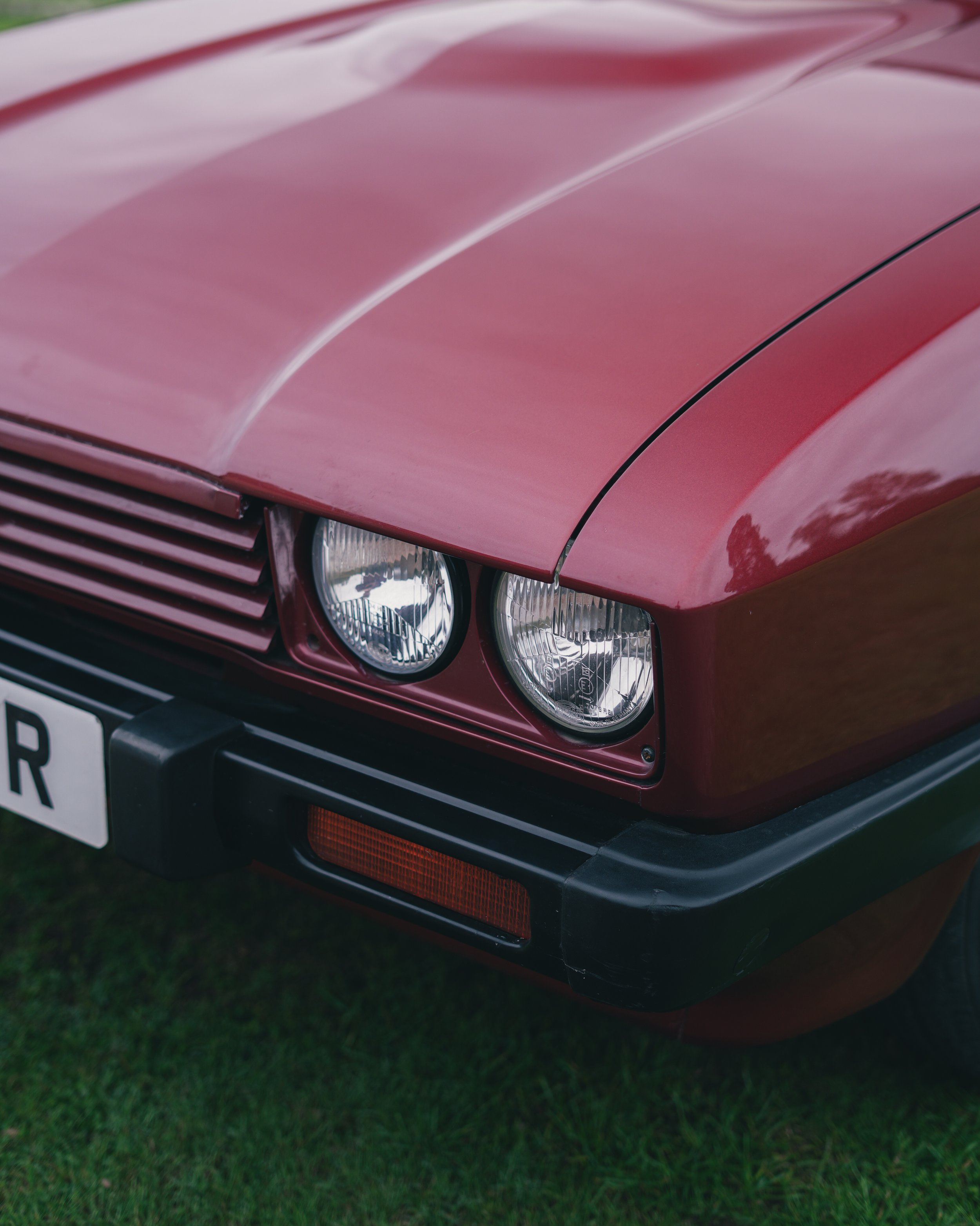 Close-up of the front right corner of a vintage red Ford Capri car showing dual headlights, part of the grille, and a black bumper with a red reflector, parked on grass.