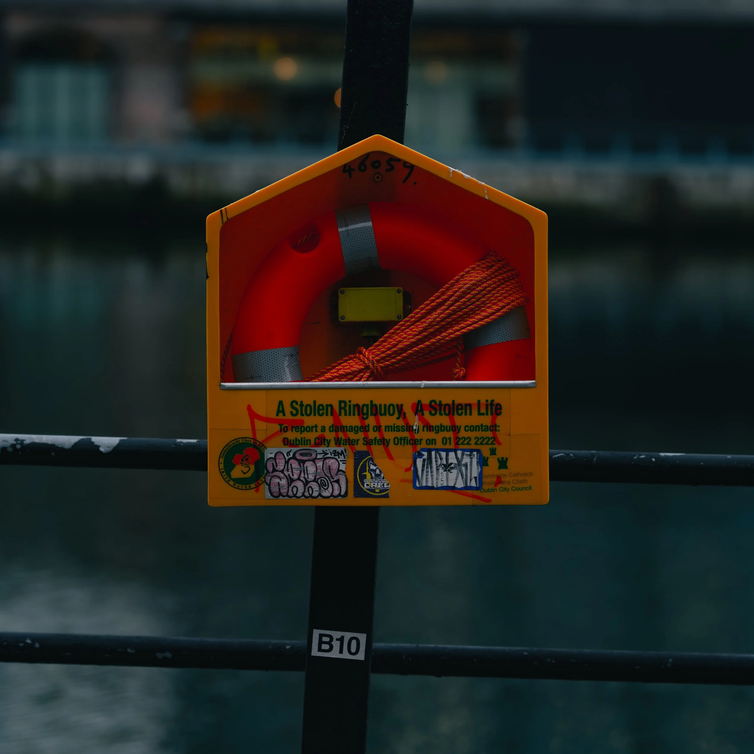 A bright yellow rescue station containing an orange lifebuoy, a folded red rescue rope, and a sticker warning about a stolen ring buoy, located on a waterfront with buildings in the background.