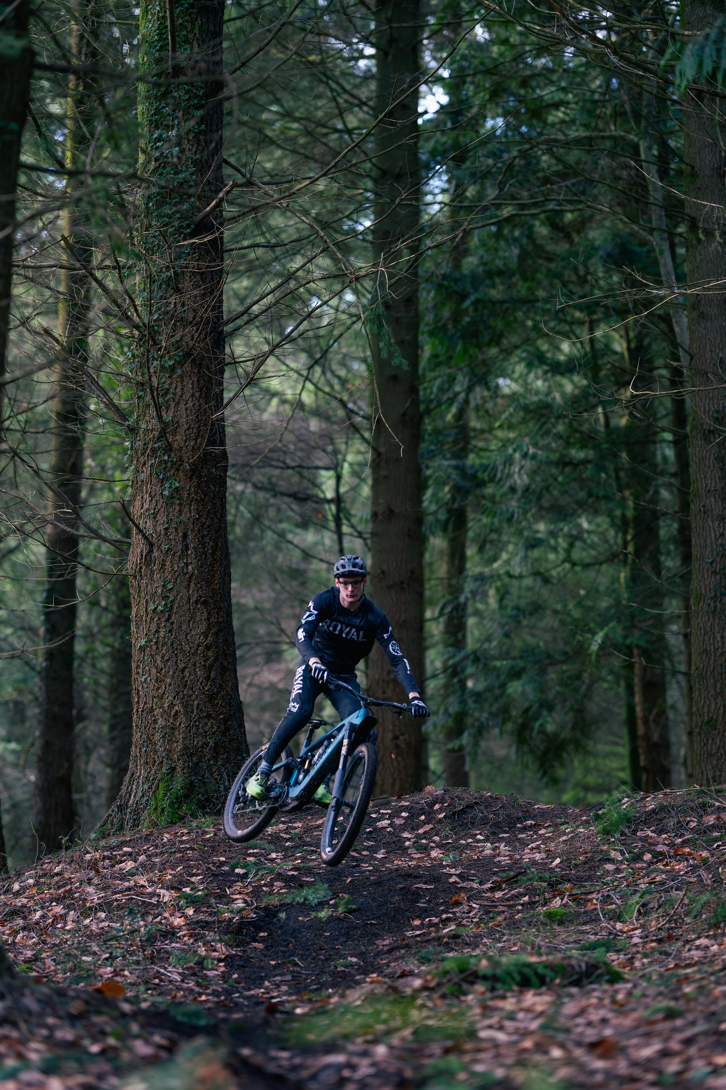 A person riding a mountain bike through a wooded forest trail surrounded by tall trees and fallen leaves.