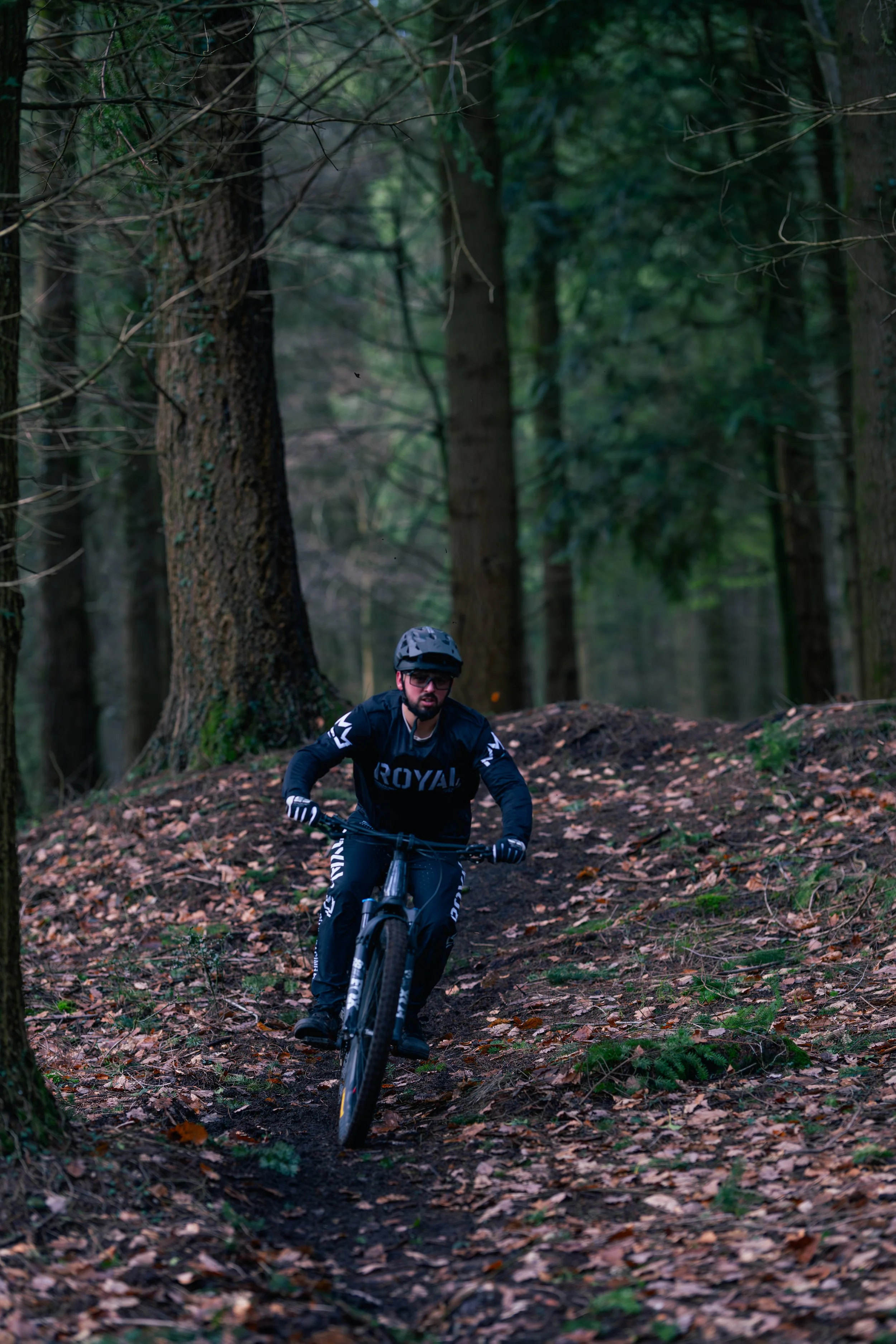 A person mountain biking on a dirt trail in a forest with tall trees and fallen leaves.