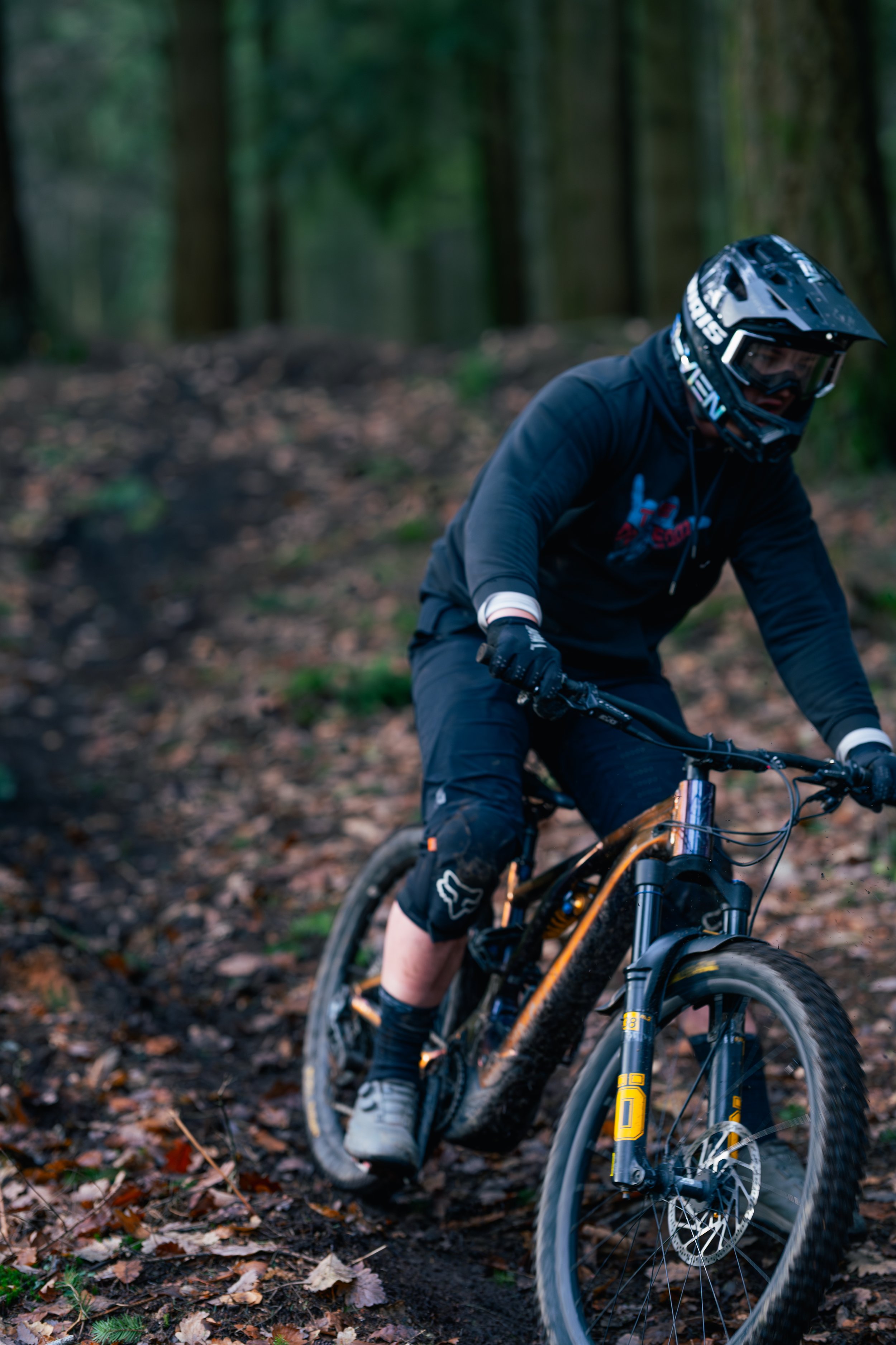 A mountain biker wearing a helmet, gloves, and black clothing riding a bike on a dirt trail in a forest.
