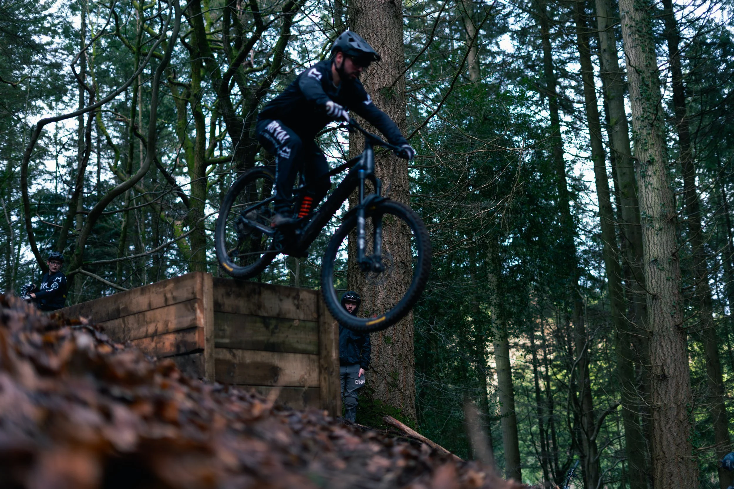 A person riding a mountain bike off a wooden ramp in a forest, with three other individuals watching nearby.