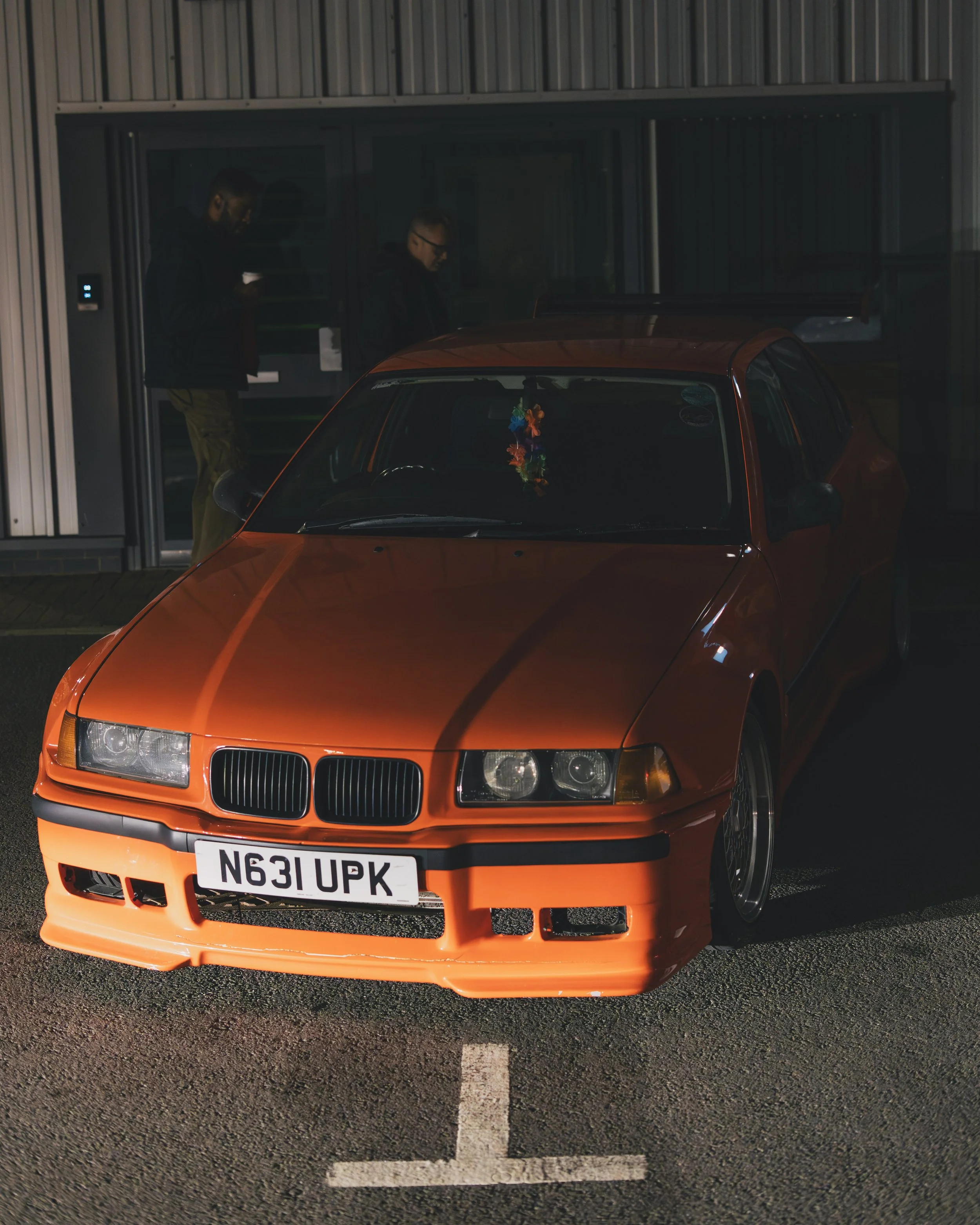 Orange sports car parked in a lot at night with two people standing in the background near a building entrance.