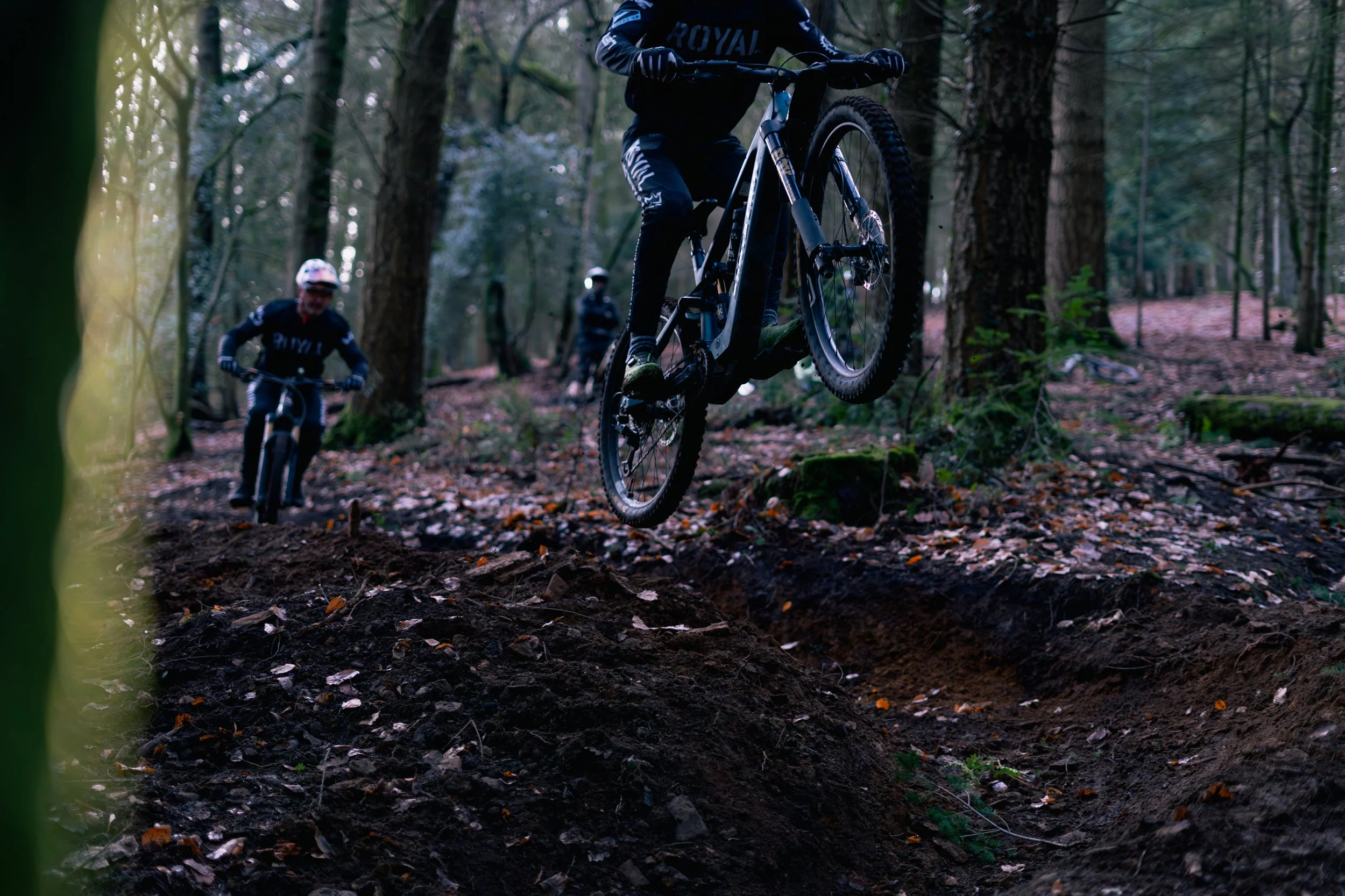 Mountain bikers riding through a wooded trail, with one cyclist in mid-air jumping over rough terrain and others following behind, in a dense forest setting.