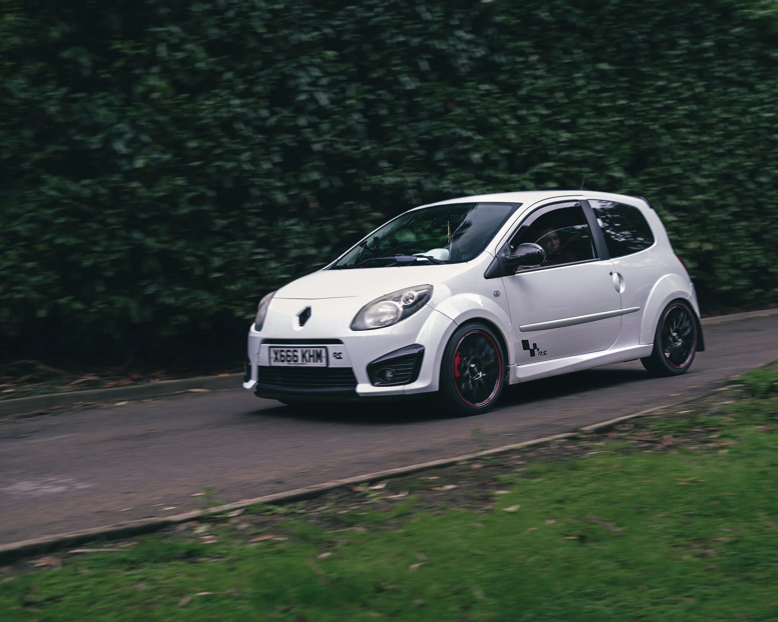 A white compact car with black wheels and red accents driving on a road near grass and bushes.