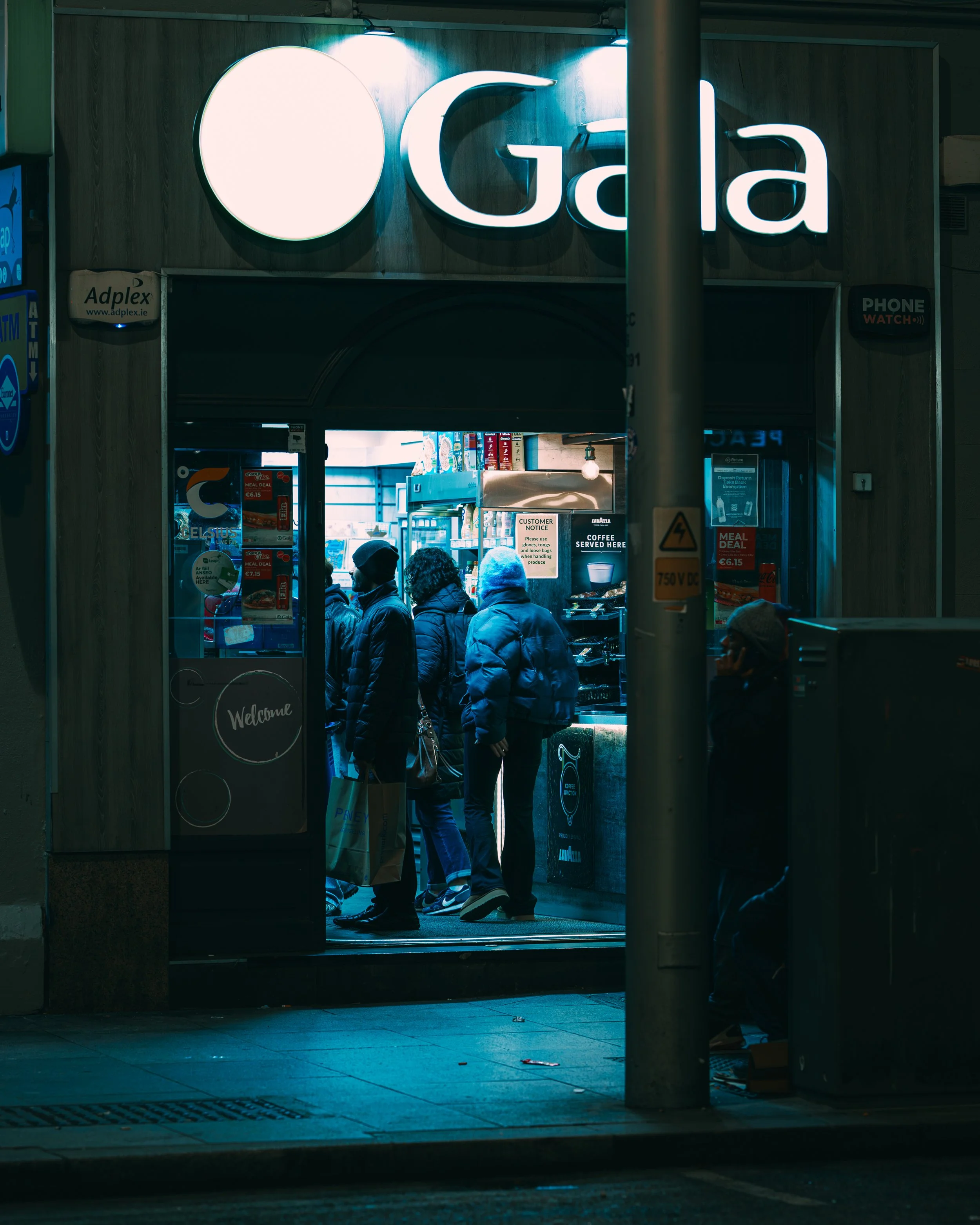 People standing in line outside a convenience store at night, illuminated by store lights, with a sign that reads 'Gala' above the entrance.