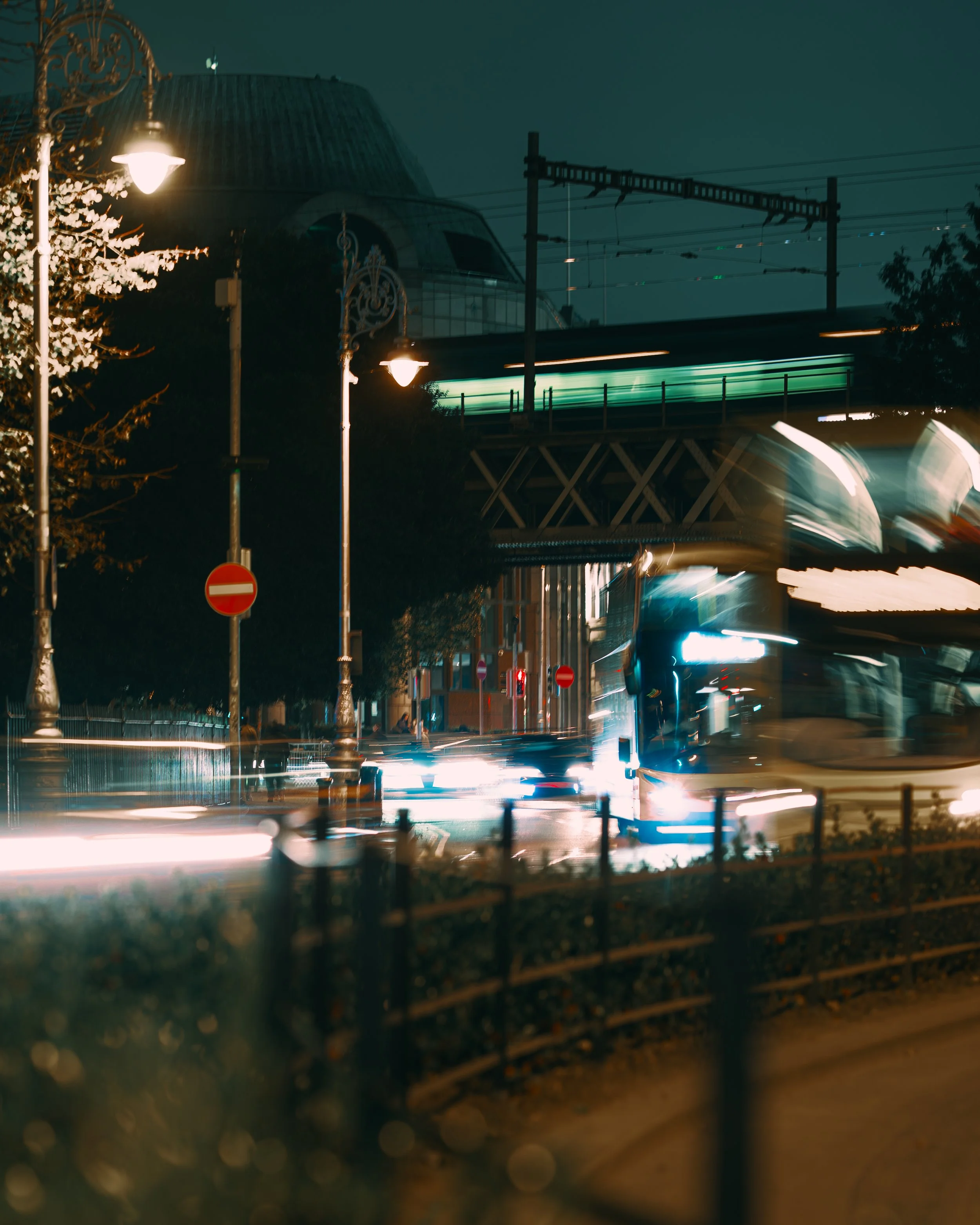 Nighttime city street with moving cars, blurred lights, street lamps, trees, and a bridge overhead.