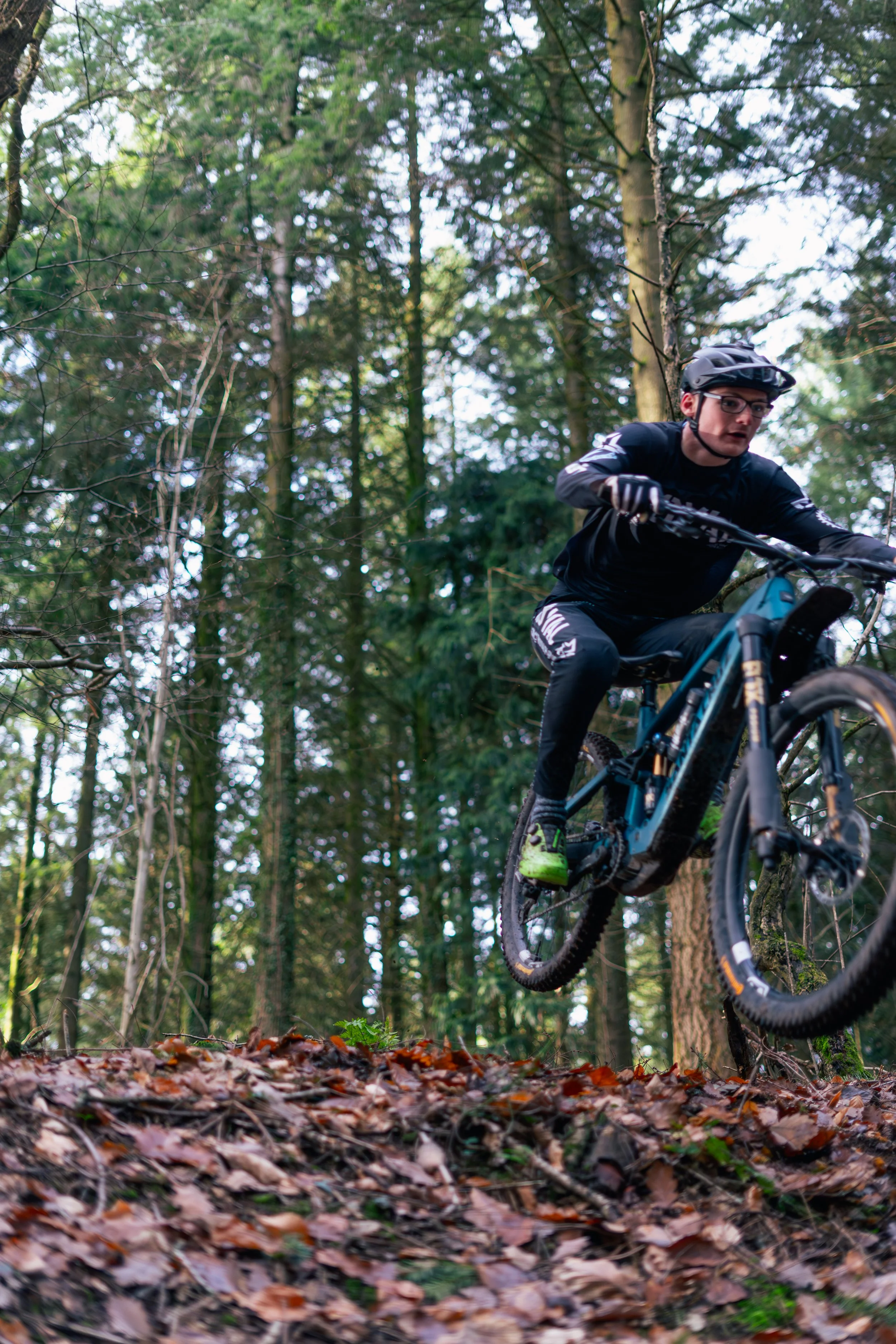A mountain biker wearing a helmet, glasses, and black sportswear riding a blue mountain bike through a forest trail covered with leaves.