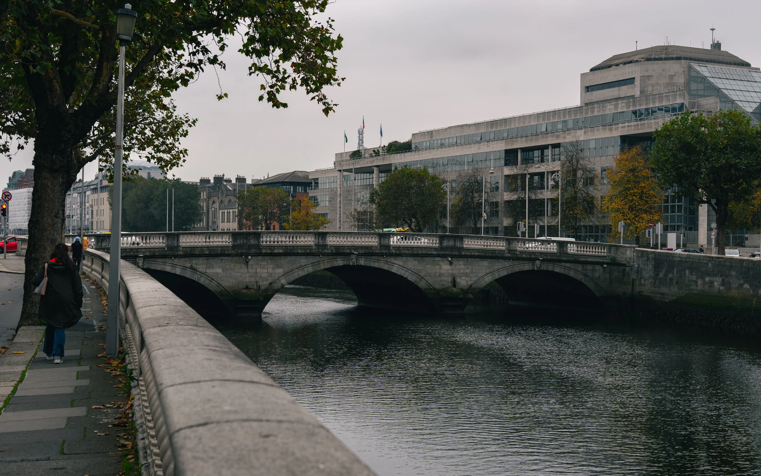 A cityscape featuring a stone bridge over a river, with a sidewalk on one side lined with trees and pedestrians, and modern buildings in the background under a cloudy sky.