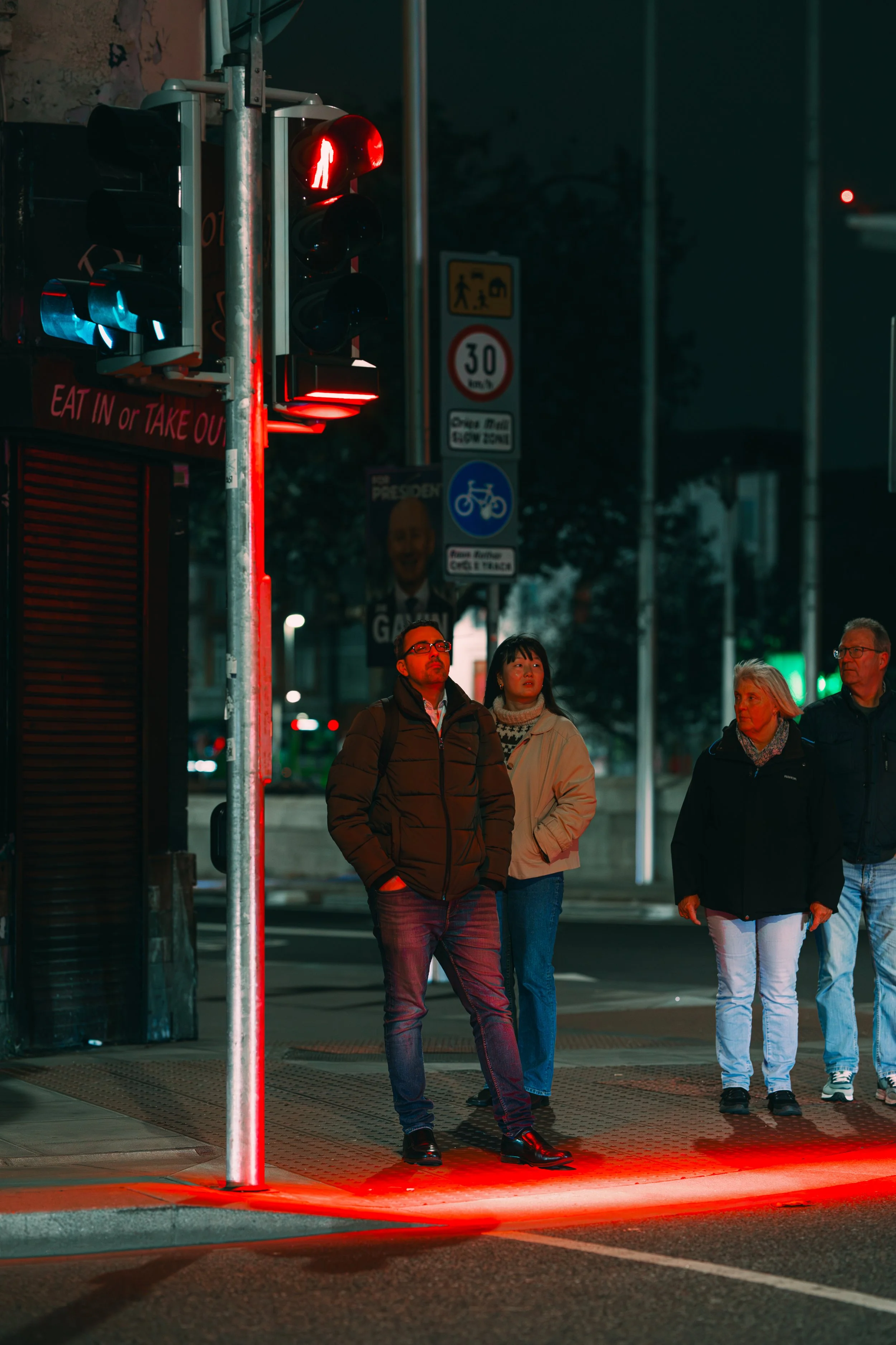 People waiting to cross at a traffic light showing a red pedestrian signal at night.