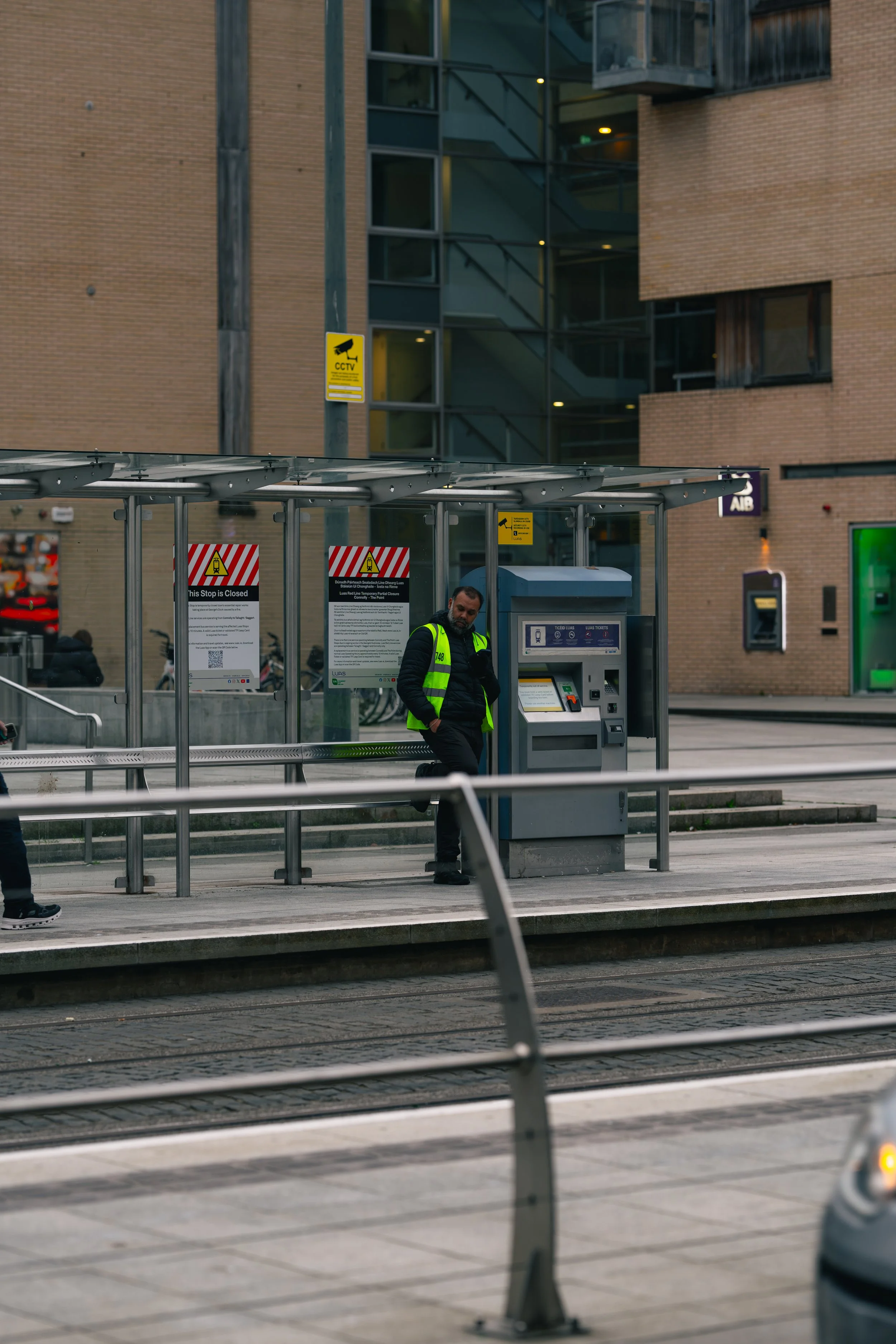 A man wearing a yellow high-visibility vest standing at a tram stop with a ticket machine, surrounded by signs and a shelter, with modern buildings in the background.