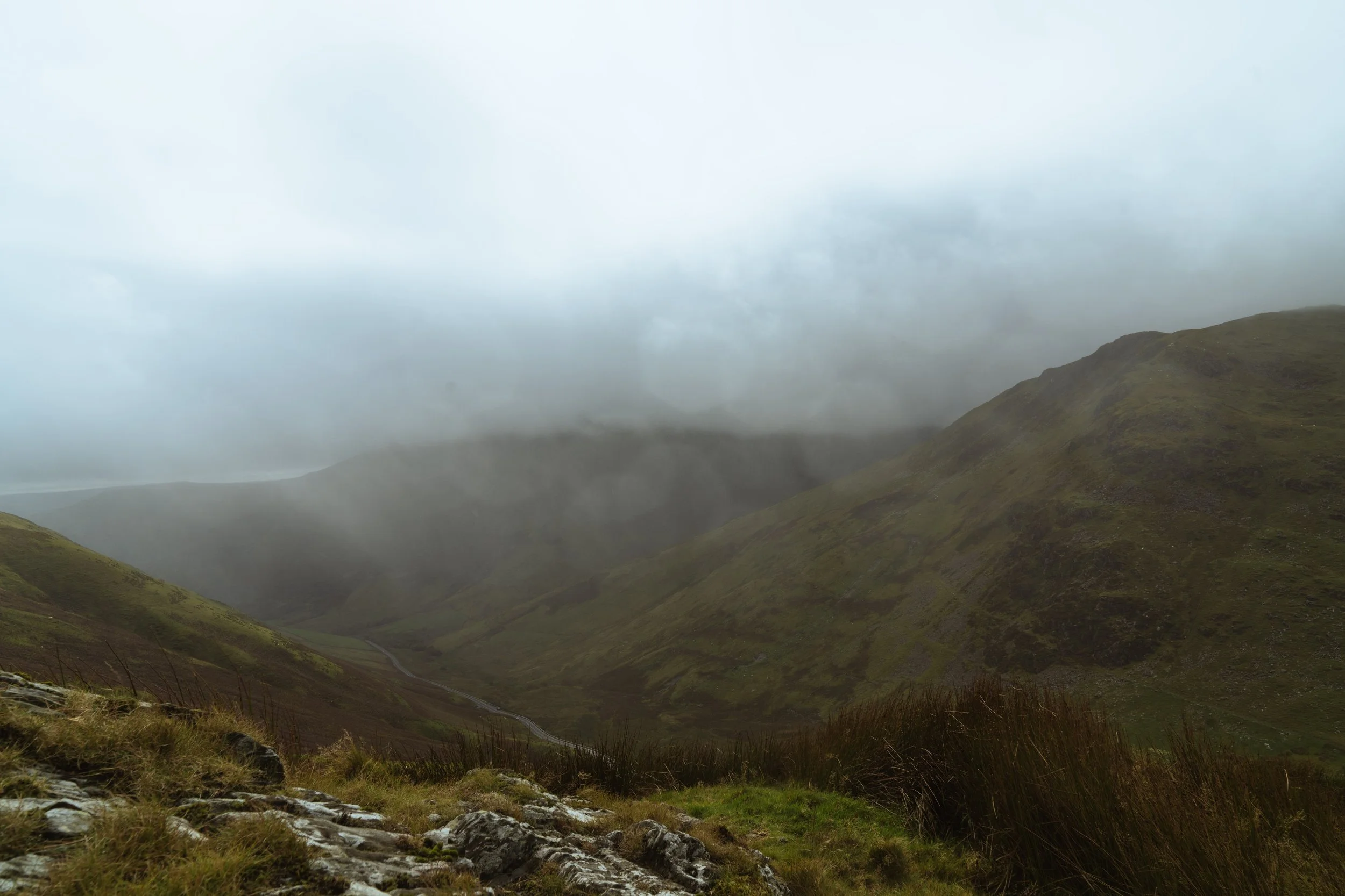 A misty mountain valley with green hills, cloudy sky, and patches of grass and rocks in the foreground. Snowdonia
