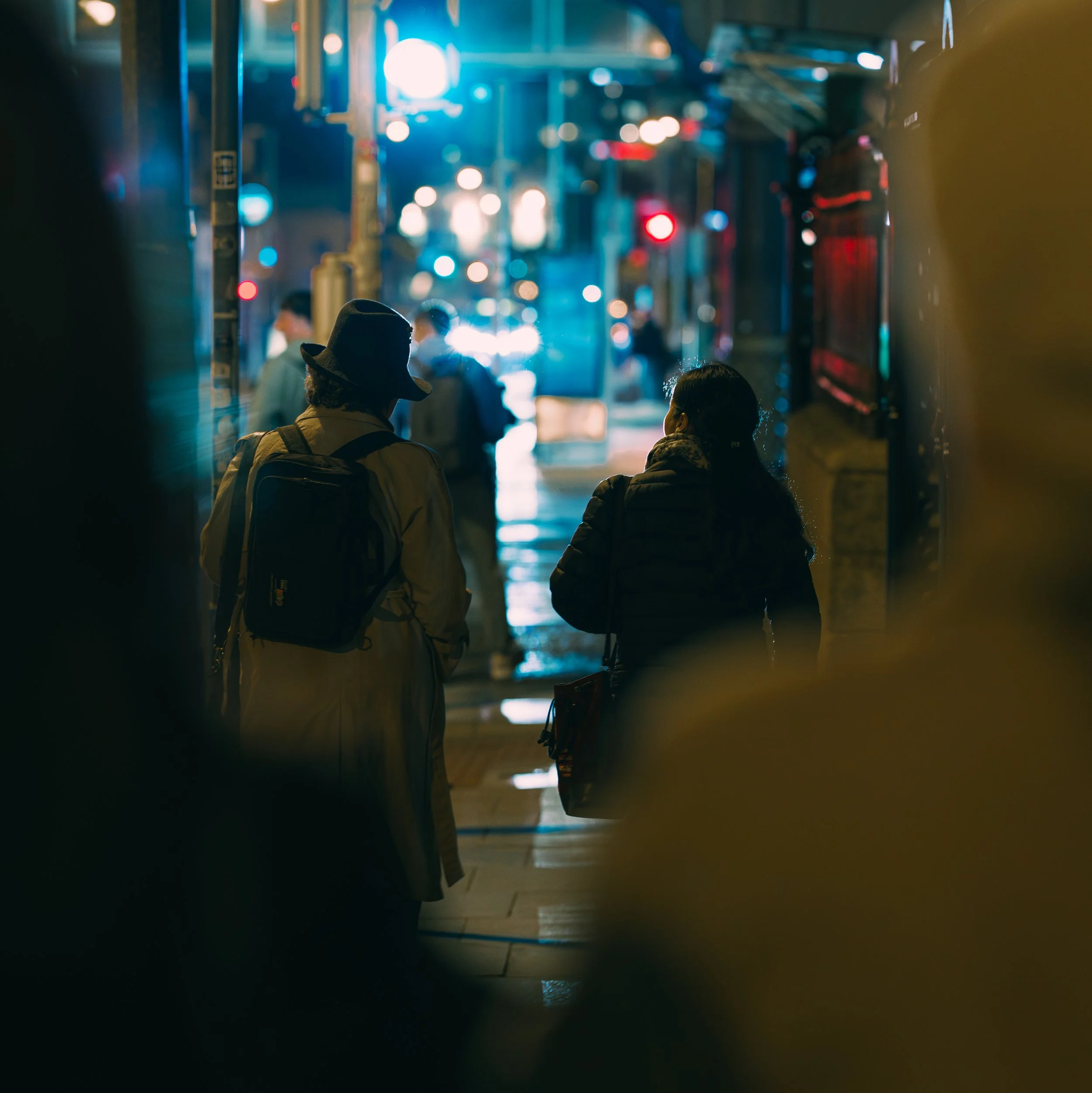 Nighttime street scene with silhouetted pedestrians walking, wet road reflecting city lights, in an urban area with neon signs and traffic.