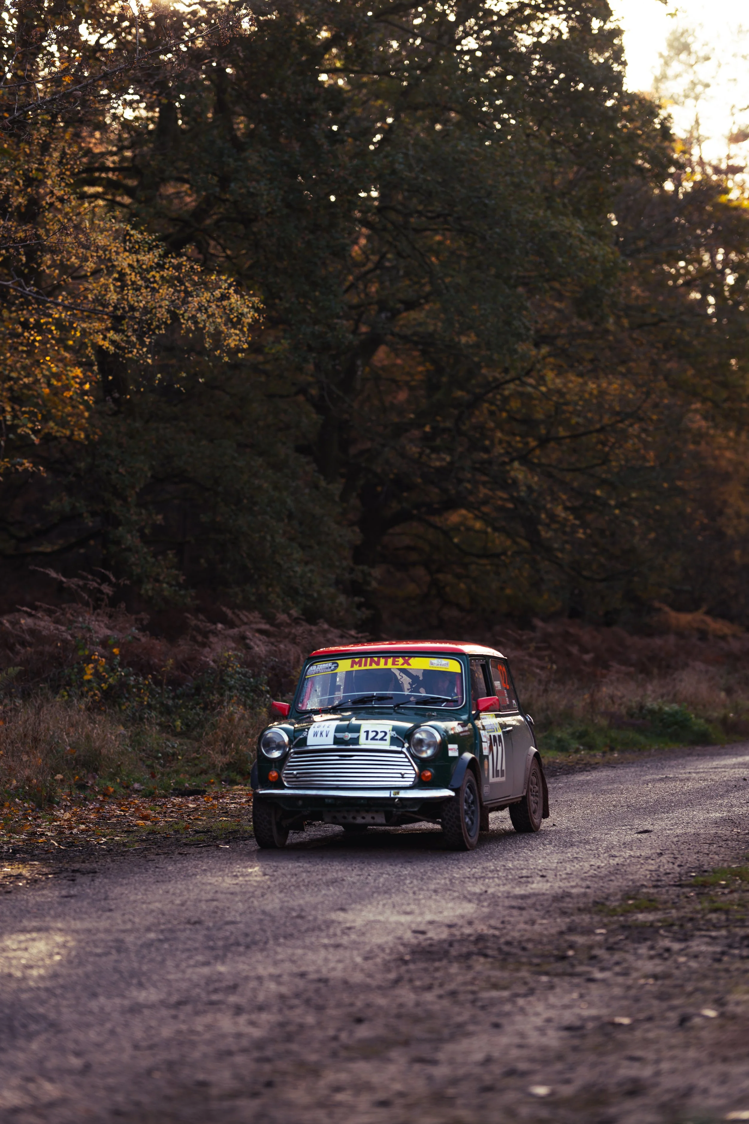 A vintage Mini Cooper race car with racing decals and the number 122, driving on a gravel road surrounded by trees during sunset. wye dean rally 