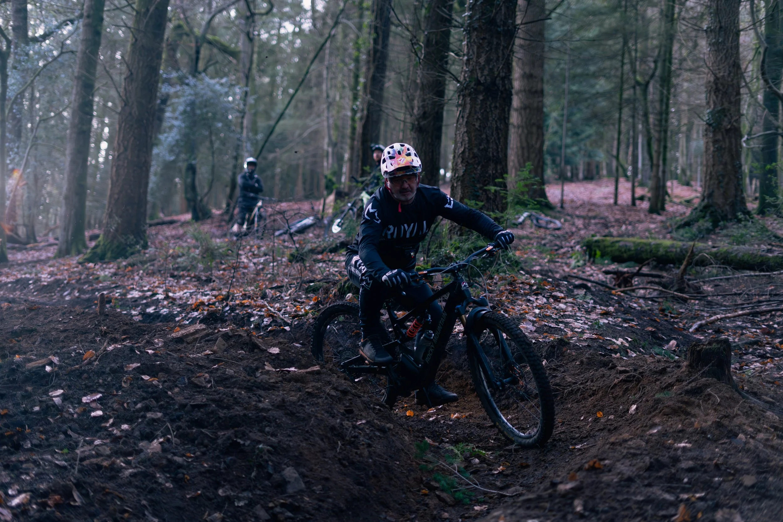 A person mountain biking on a dirt trail in a forest, wearing a helmet and sports gear, with two other bikers visible in the background.
