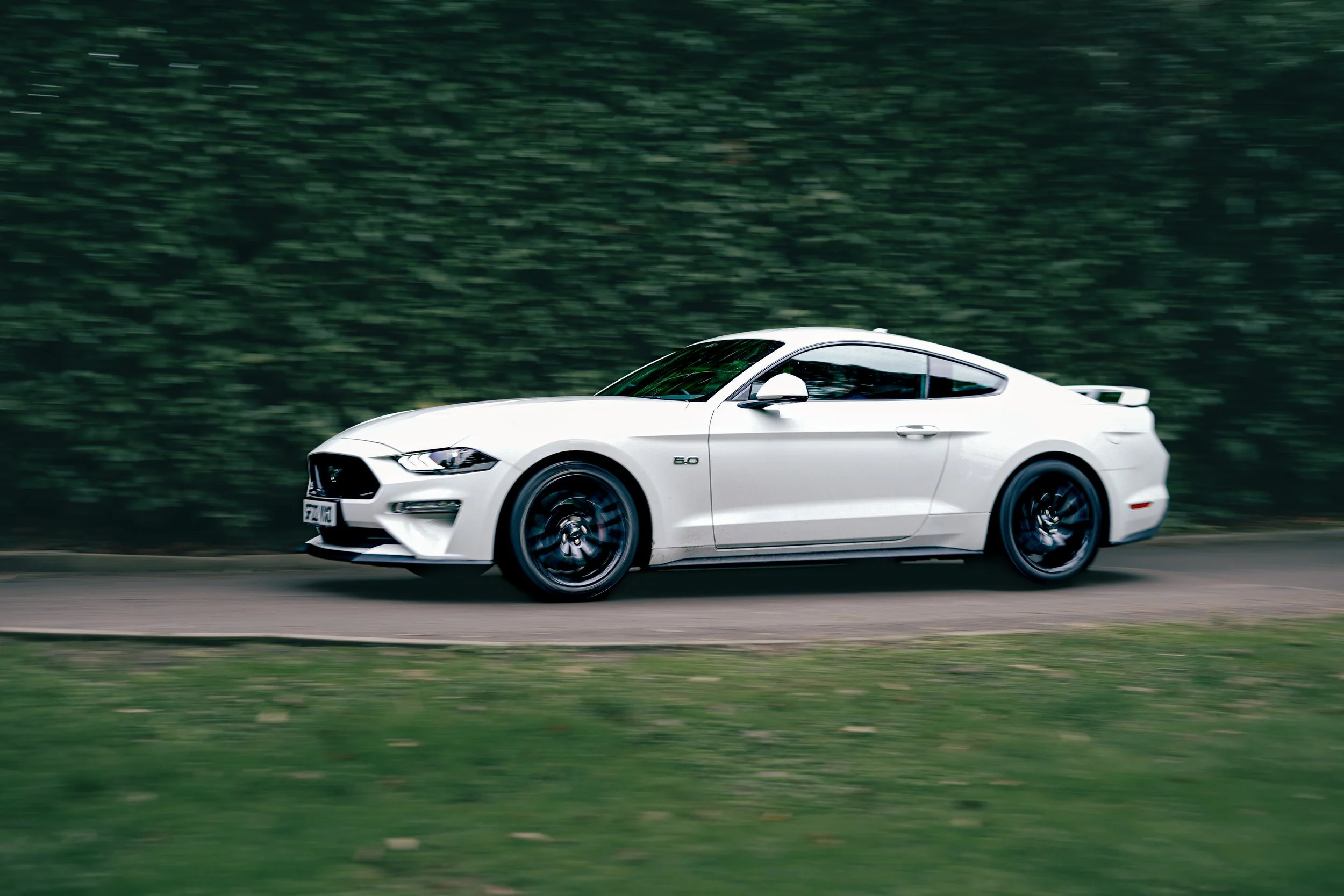 A white Ford Mustang sports car driving on a road with a green, blurred background.