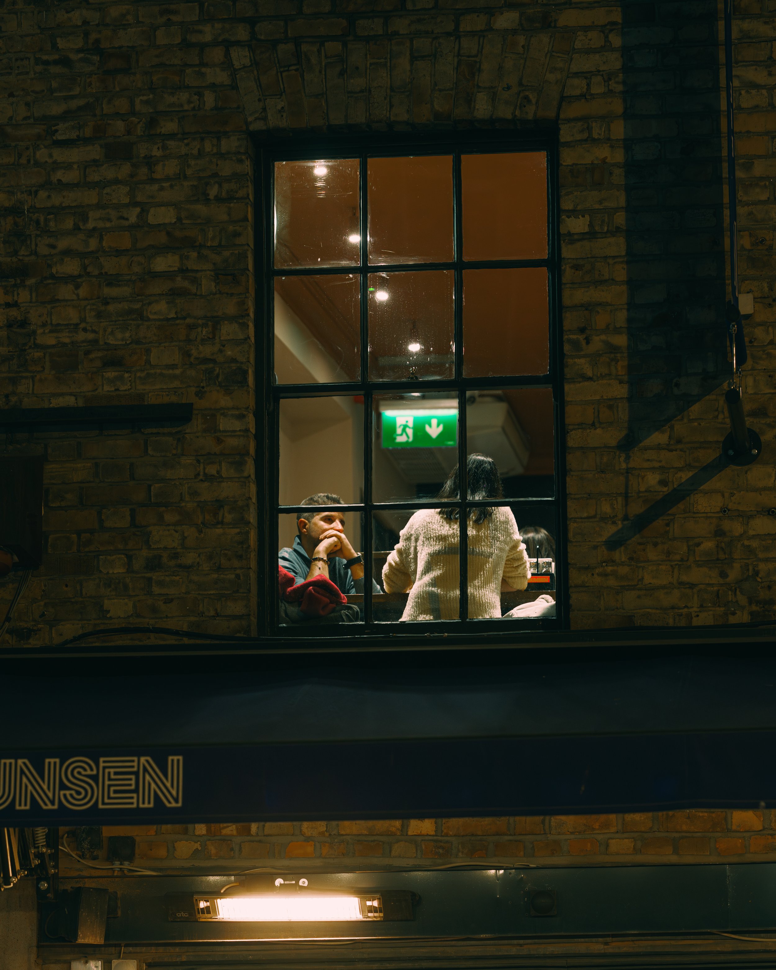 A view through a window showing two people sitting at a table inside a restaurant or café, with the brick wall exterior and a green emergency exit sign visible inside.