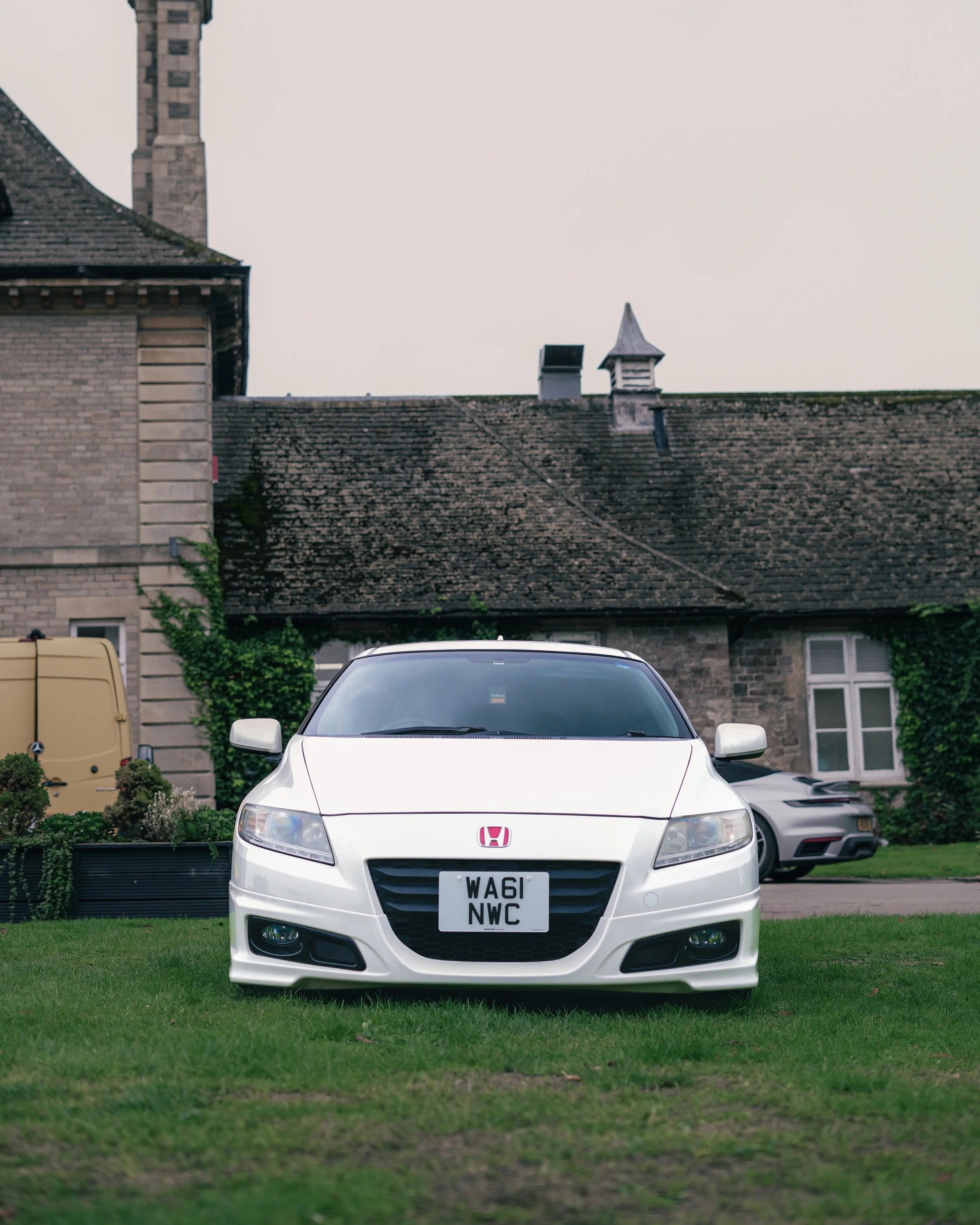 Front view of a white Honda CR-Z car parked on a grassy lawn with a house and another vehicle in the background.