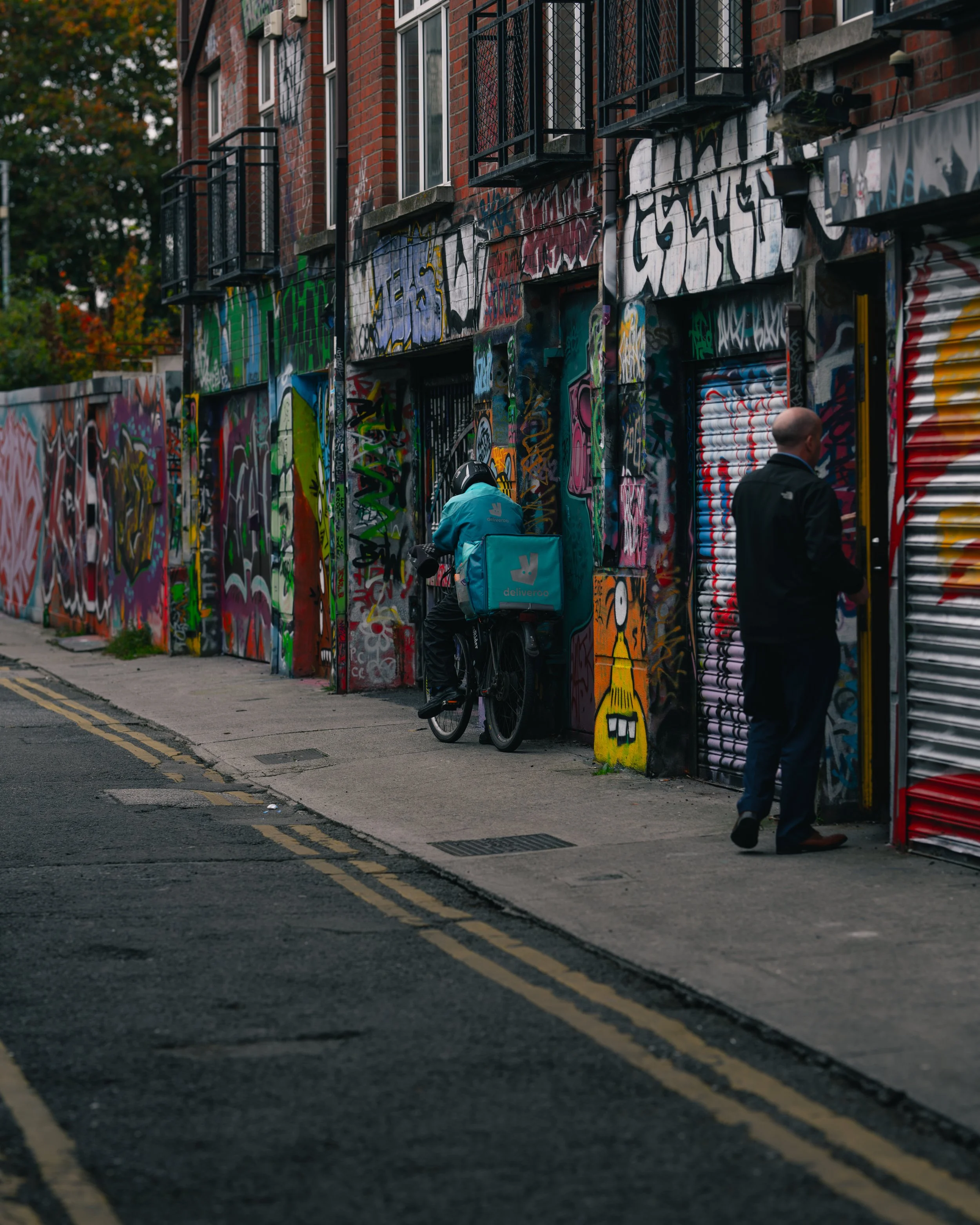 A city street with a wall covered in colorful graffiti art, a delivery person on a bicycle, and a man walking past a closed storefront.