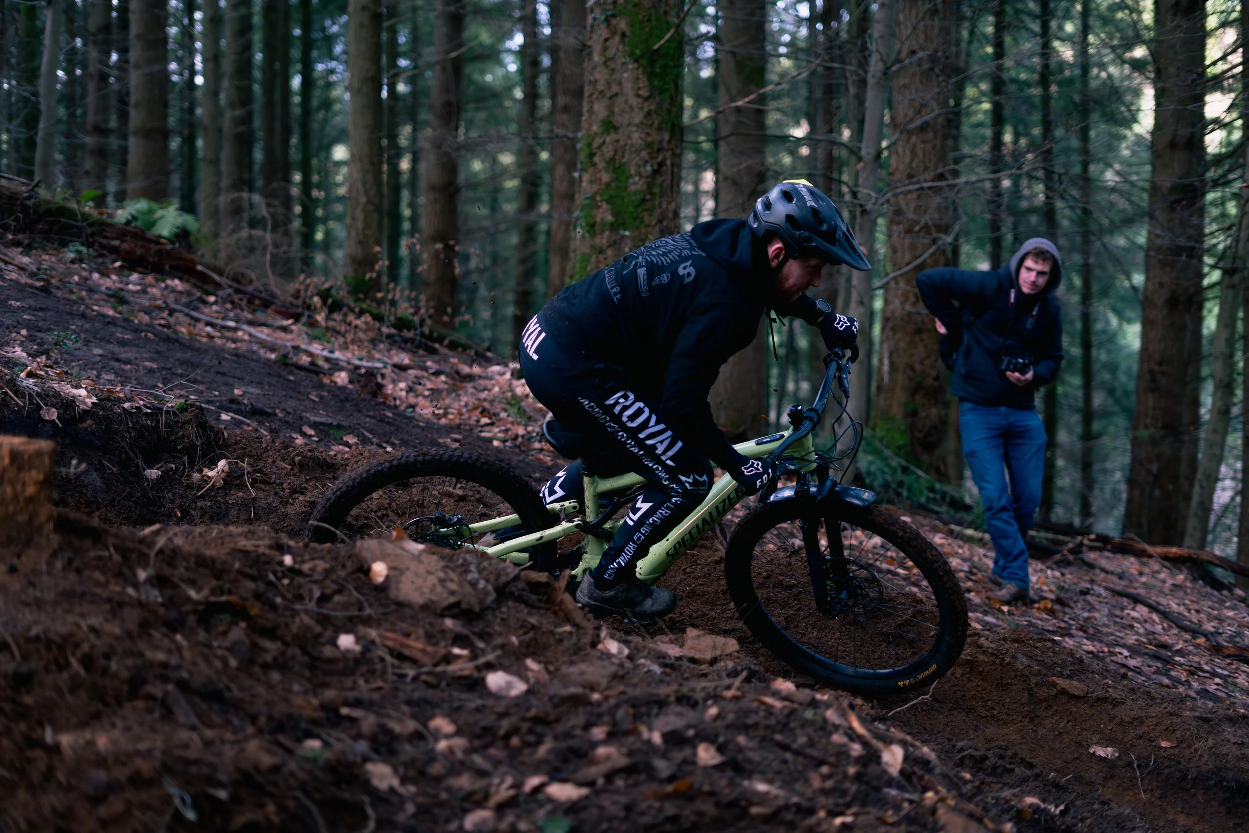 A person mountain biking downhill on a forest trail with another person observing in the background.