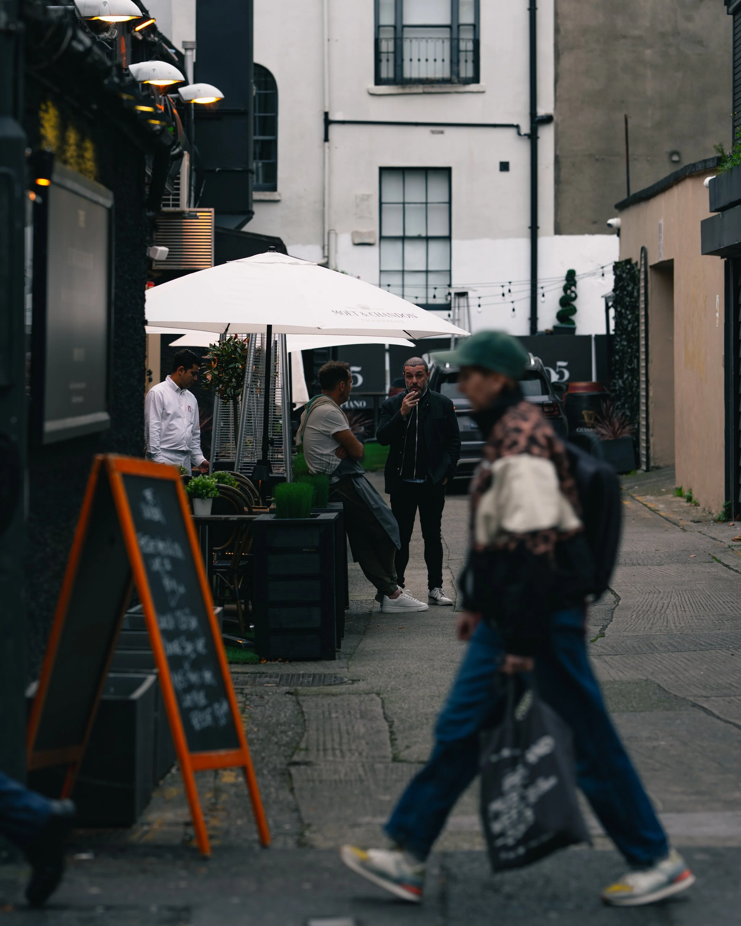 People walking and talking outside a café with outdoor seating and a large white umbrella, in an urban setting with buildings and parked cars.