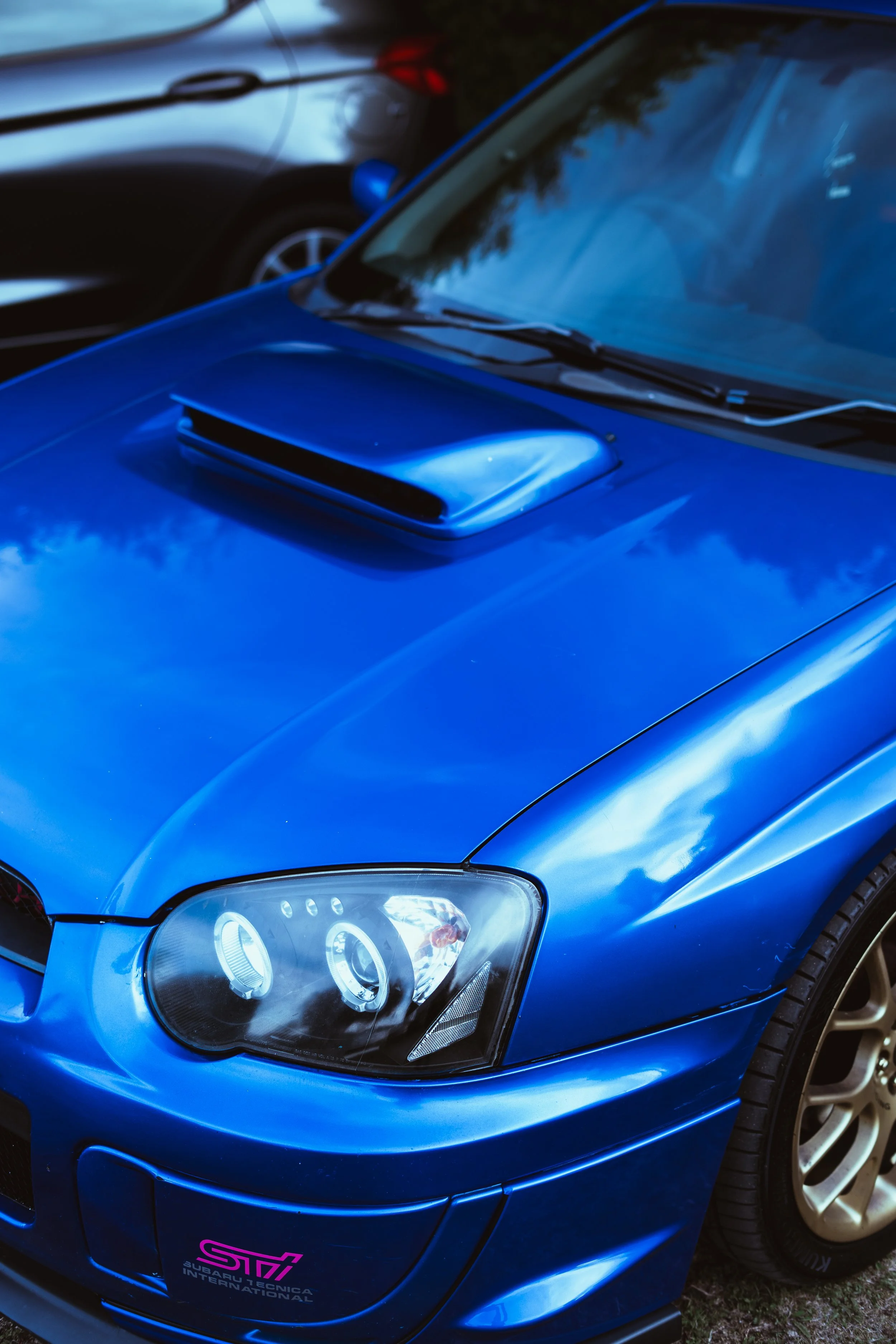 Close-up of a blue Subaru car with a prominent hood scoop and a pink Subaru logo on the front bumper, parked outdoors.
