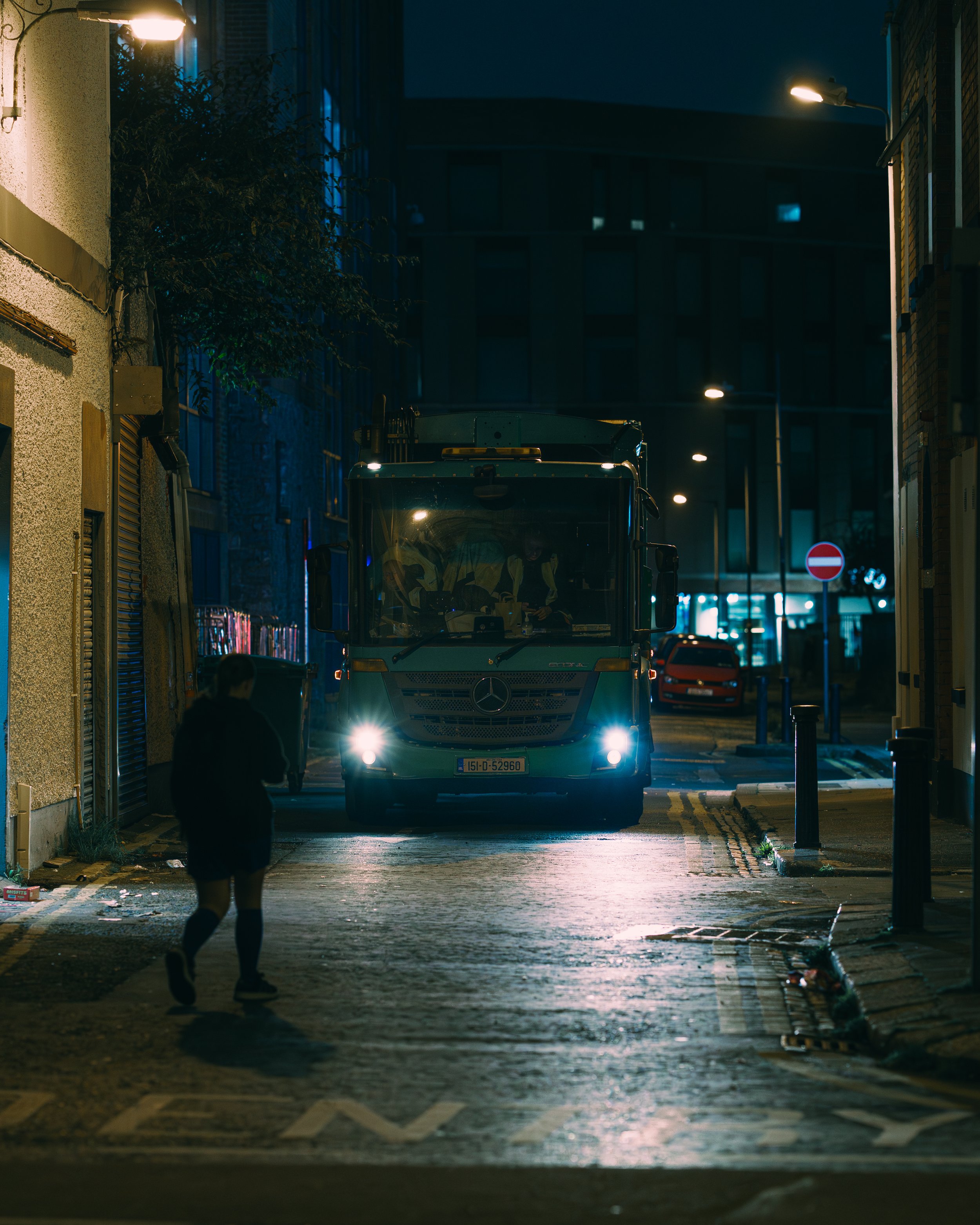 Nighttime street scene with a large truck facing the camera, headlights on, and a person walking away in the foreground.