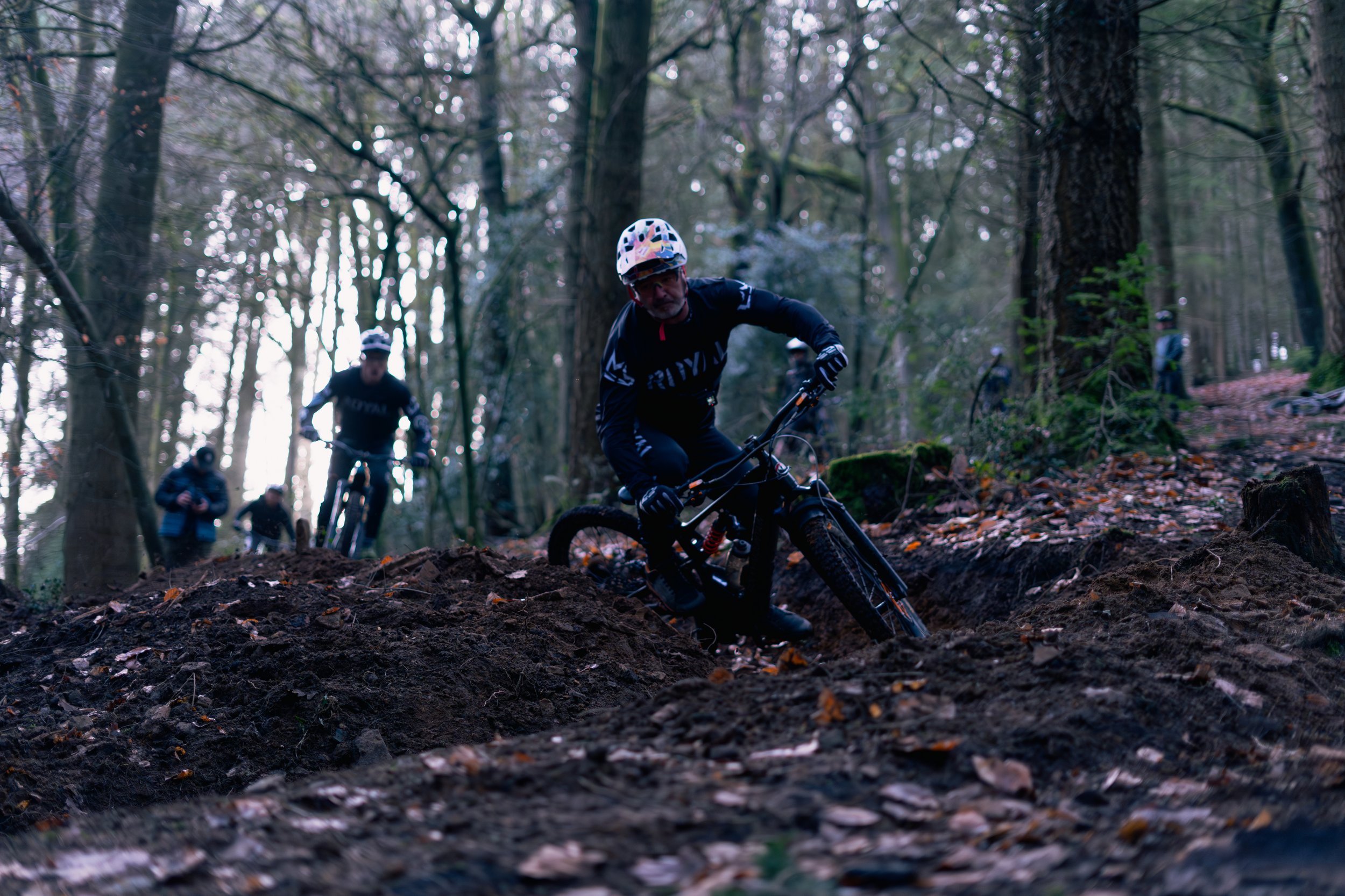 Man mountain biking downhill on a dirt trail in a forest, wearing a helmet and black riding gear, with several other bikers in the background.