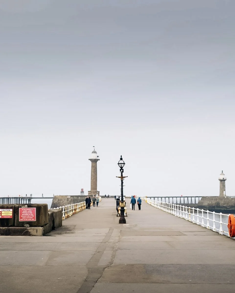 A pier extending into the water with lighthouse structures at the end and a few people walking along it on a cloudy day.