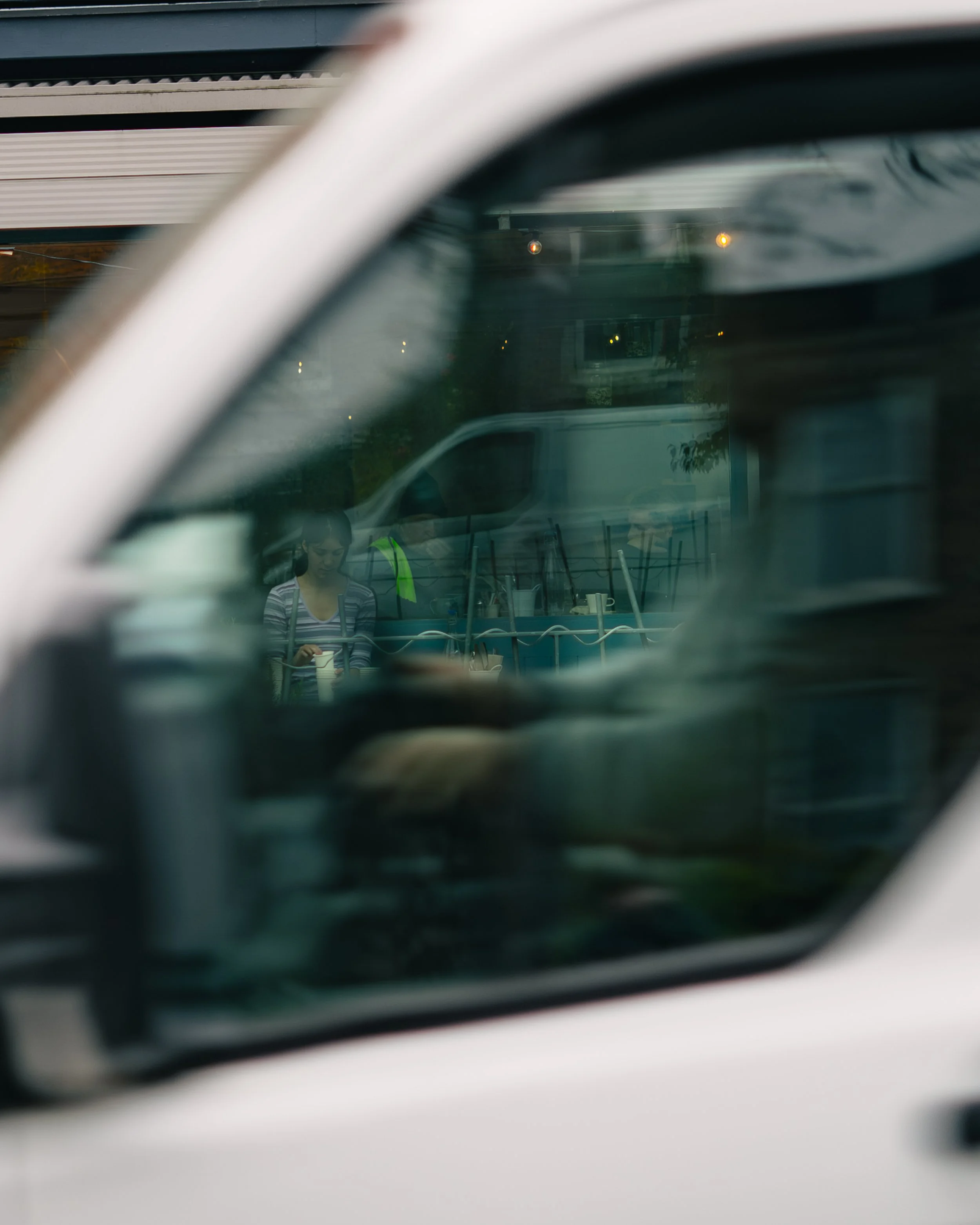 Reflected image of a woman in a striped shirt looking down, seen through a car window with other vehicles and a building in the background.