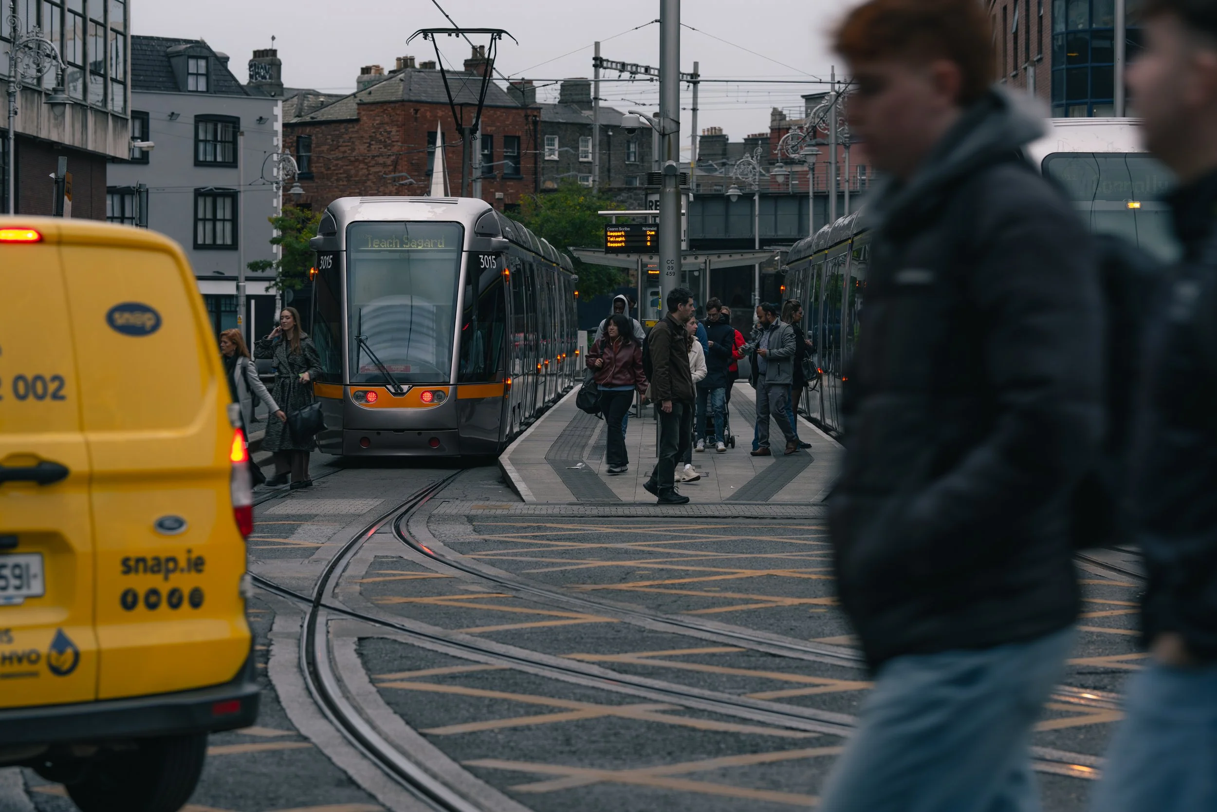 People walking at a tram stop with a modern tram approaching. There is a yellow delivery van on the road with the logo 'snap.ie' and the website address. The background shows urban buildings and electrical wires for the tram system. 