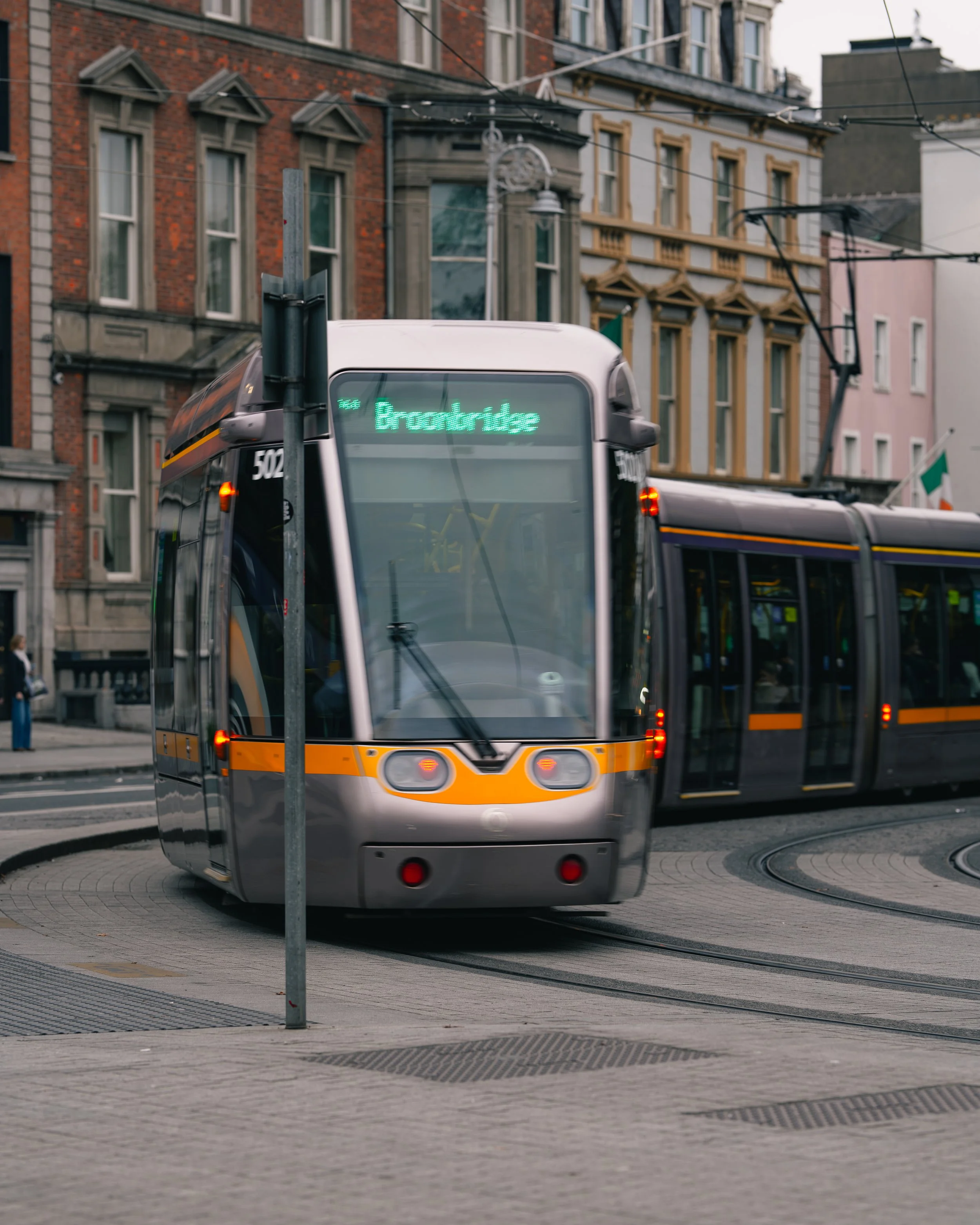 A modern light rail tram on the street with an illuminated sign indicating its destination as 'Broadway.' The tram is silver with orange and black accents, moving along curved tracks with buildings and overhead wires in the background.