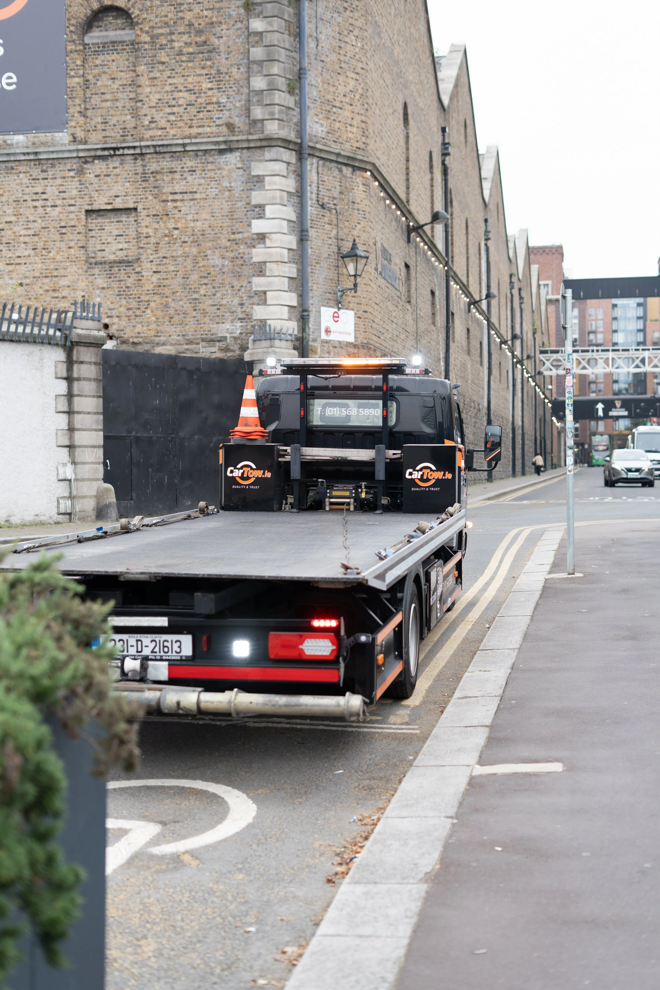 A tow truck parked next to a brick building on a city street, with traffic and pedestrians in the background.