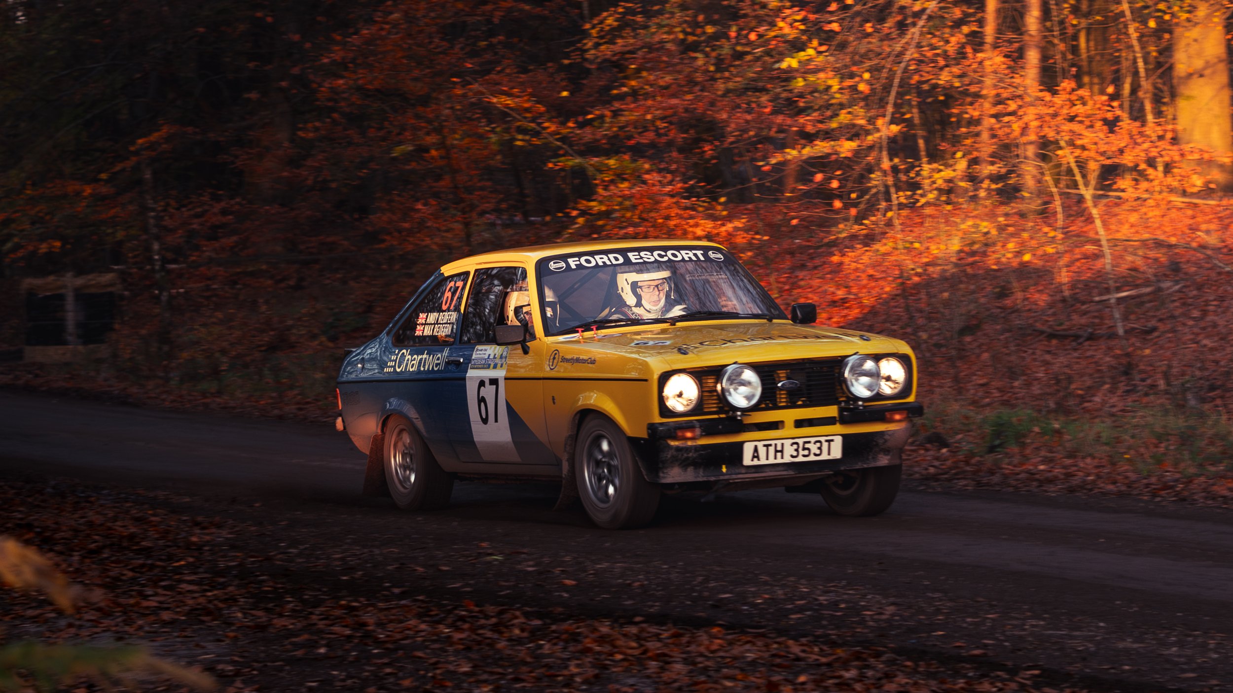 A yellow and blue vintage rally car with the number 67 racing on a dirt road surrounded by autumn foliage in a forest. wye dean rally 