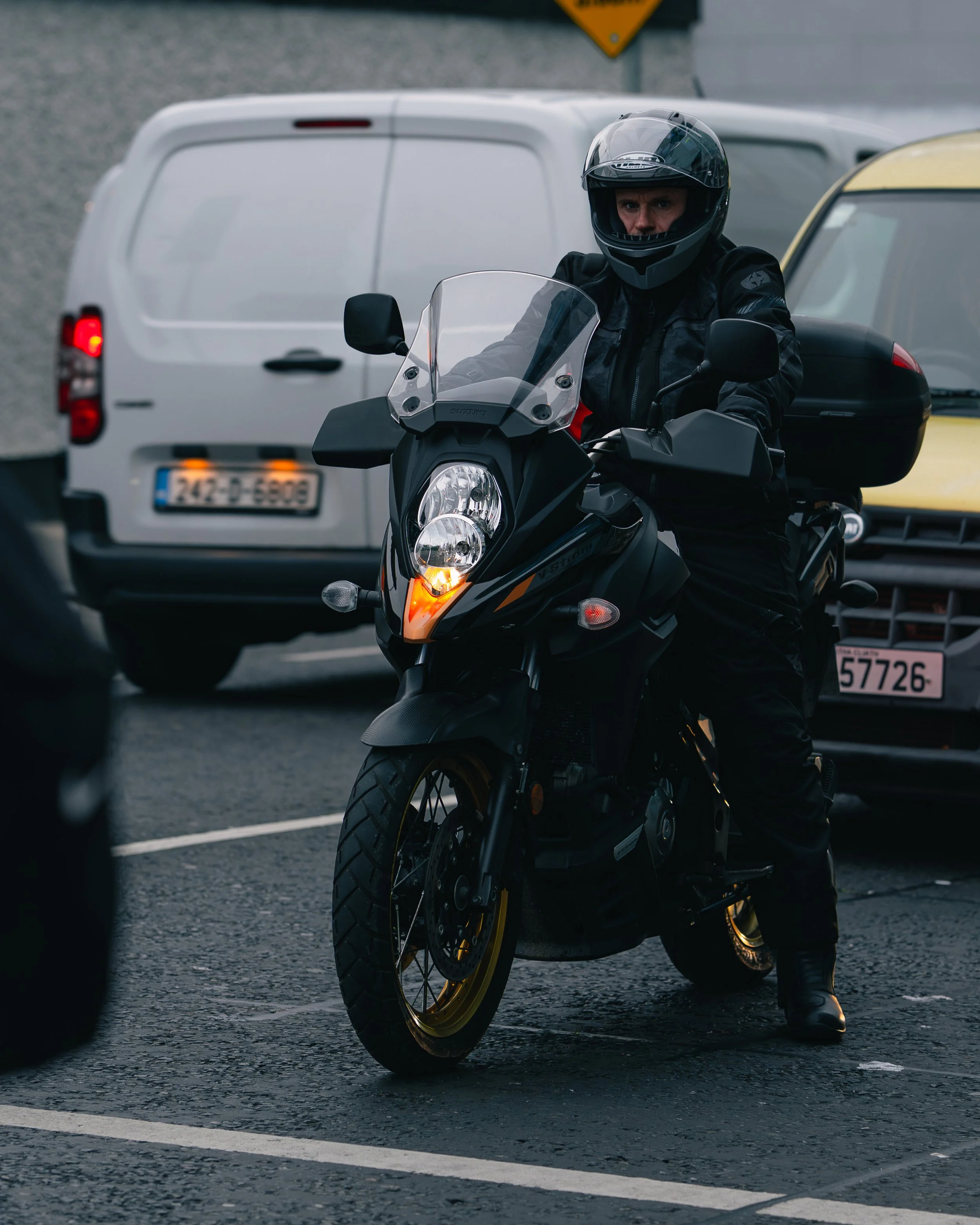 Motorcyclist wearing black gear and a helmet, sitting on a black motorcycle, in a parking lot with white and yellow cars in the background.