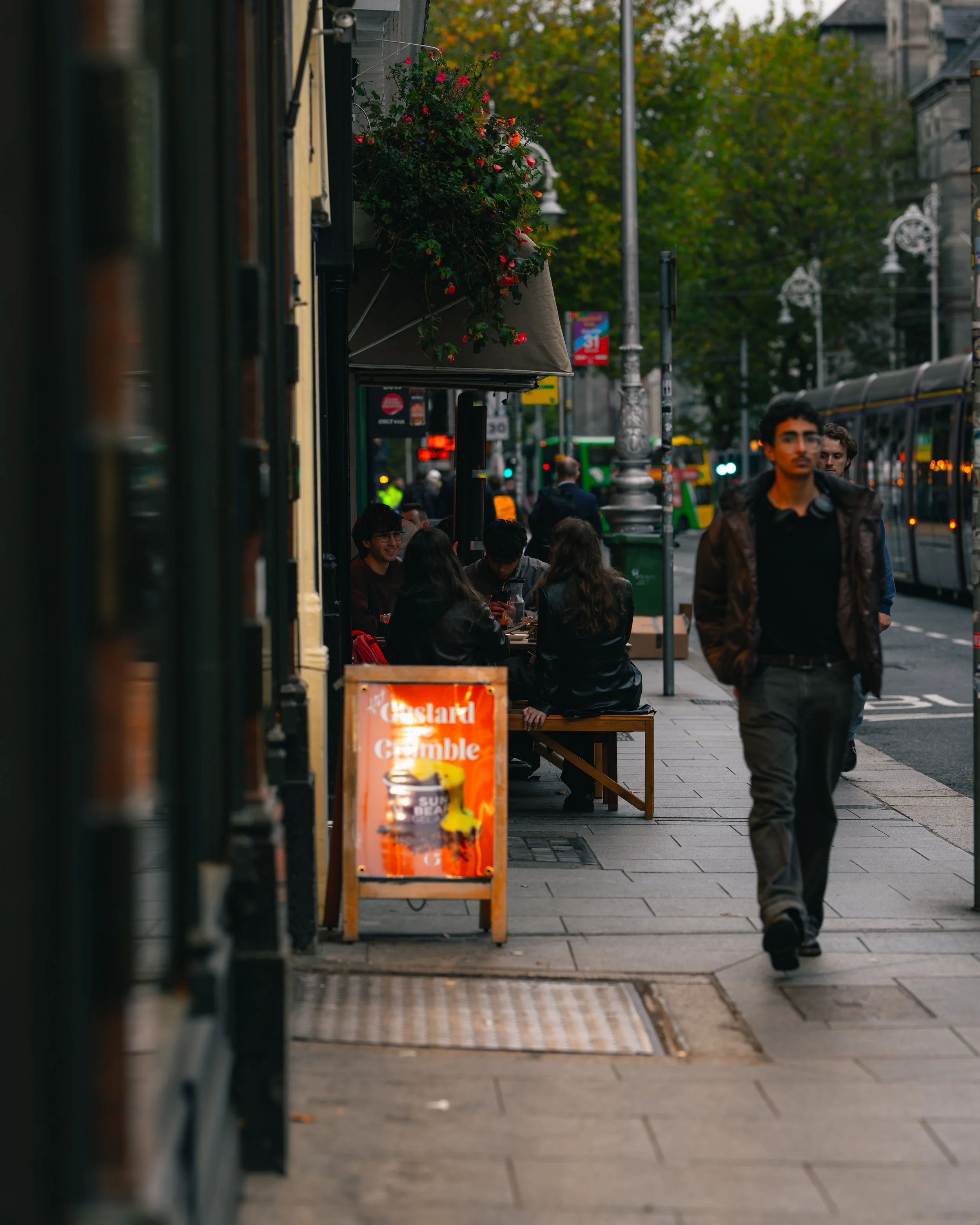 People sitting at outdoor tables on a city sidewalk, with a man walking past. A sign in the foreground reads "Hazard Gamble."