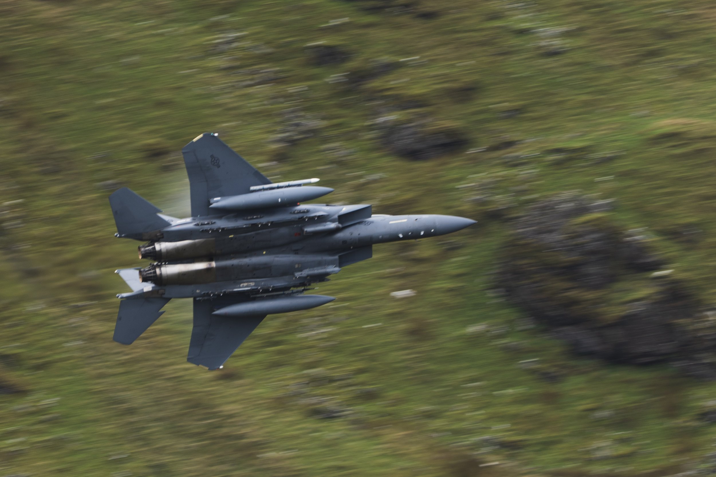 A fighter jet flying at high speed over a grassy terrain with rocks, creating a motion blur effect.