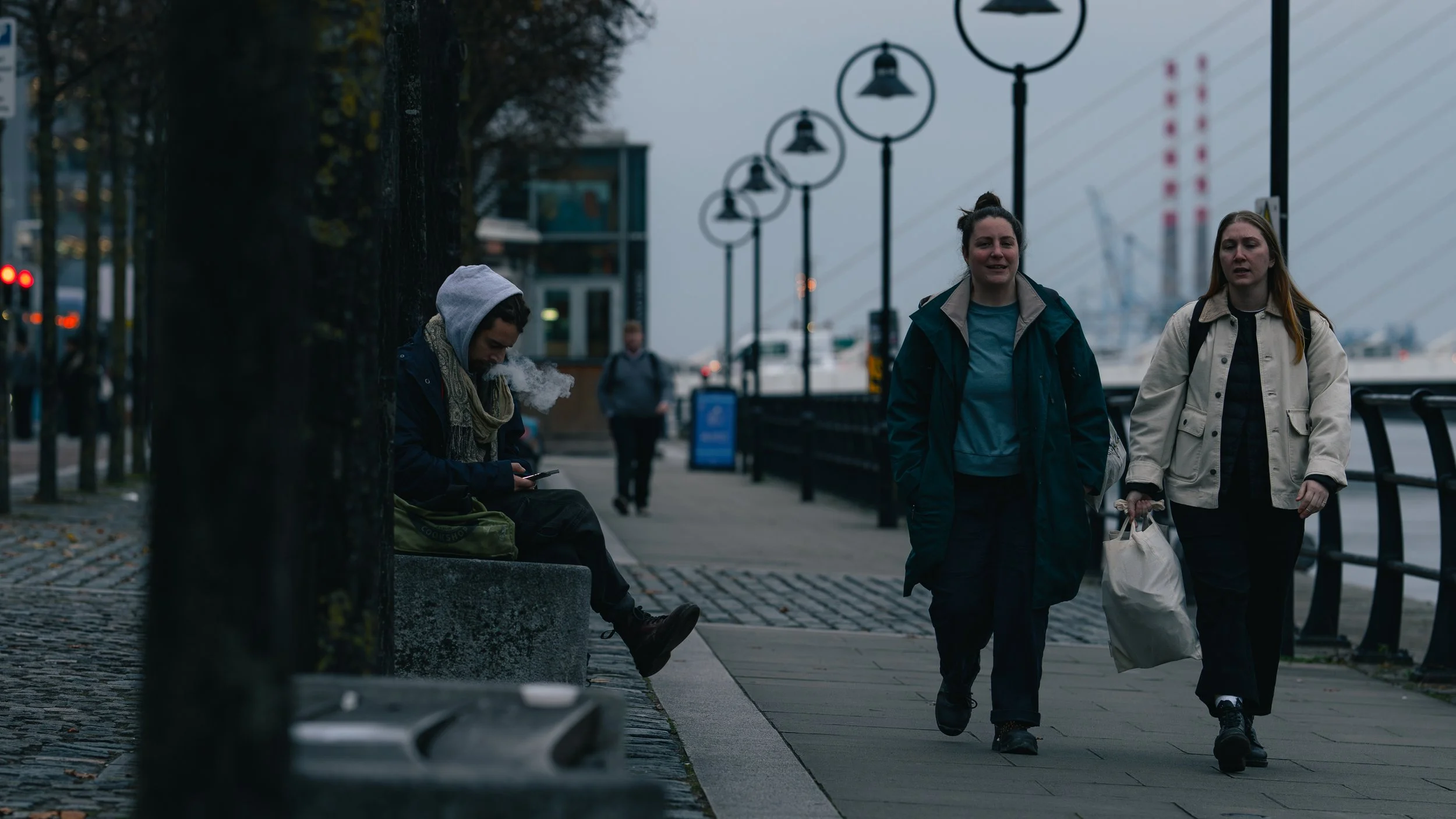 Two women are walking along a waterfront promenade during dusk, while a young man sits on a bench nearby, looking at his phone. The background features a bridge with suspension cables and street lamps.