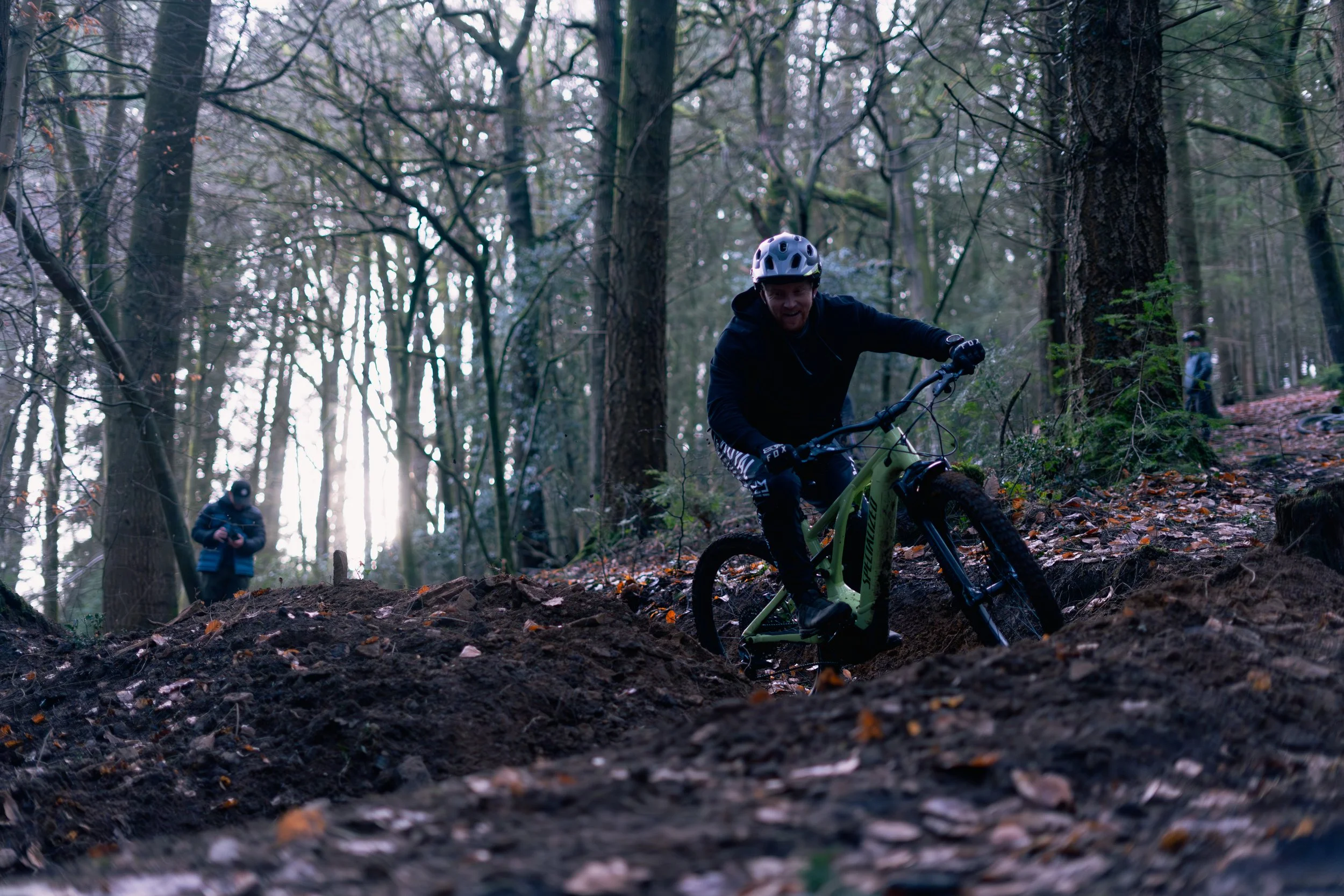 A person mountain biking downhill on a rugged trail in a forest, wearing a helmet and dark clothing, with two other people in the background among tall trees.