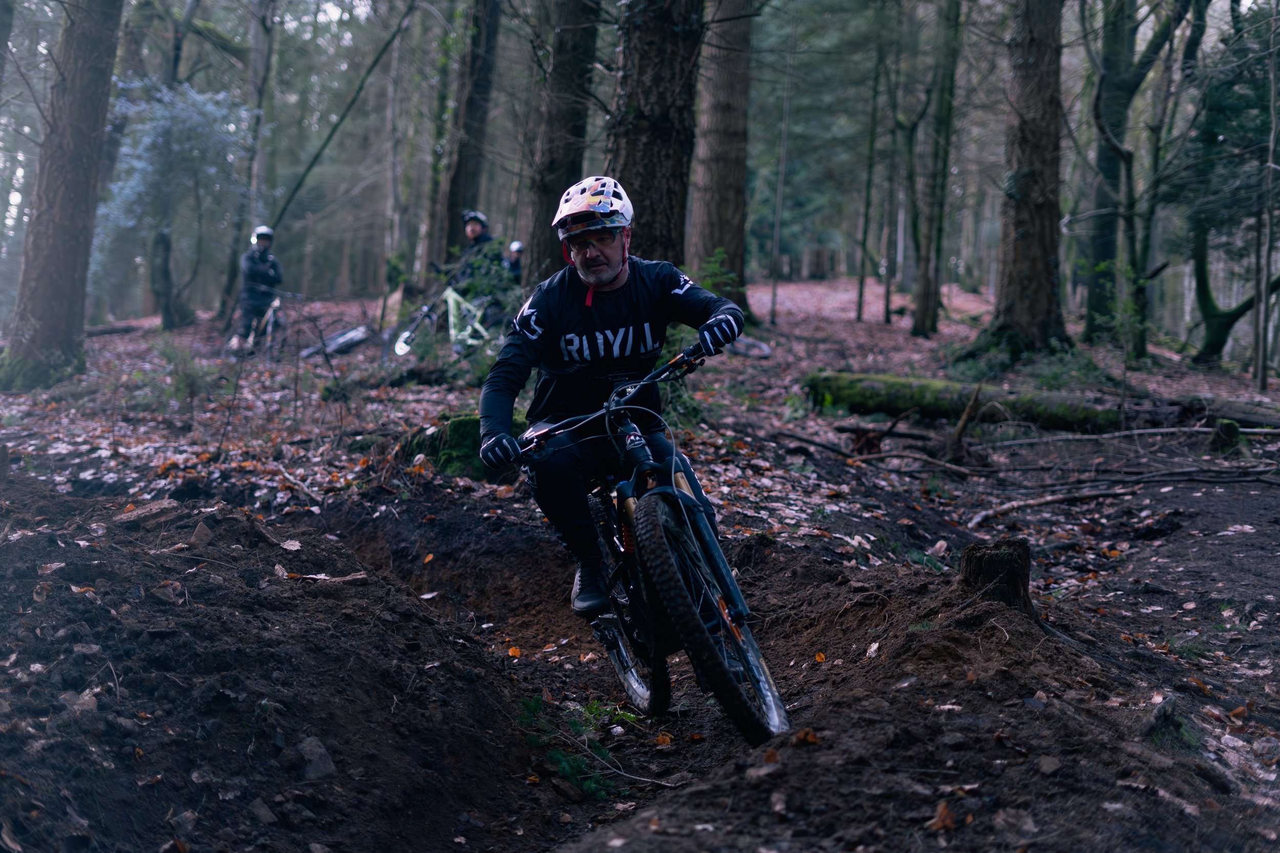 A group of mountain bikers riding on a dirt trail in a dense forest with tall trees, fallen leaves, and uneven terrain.