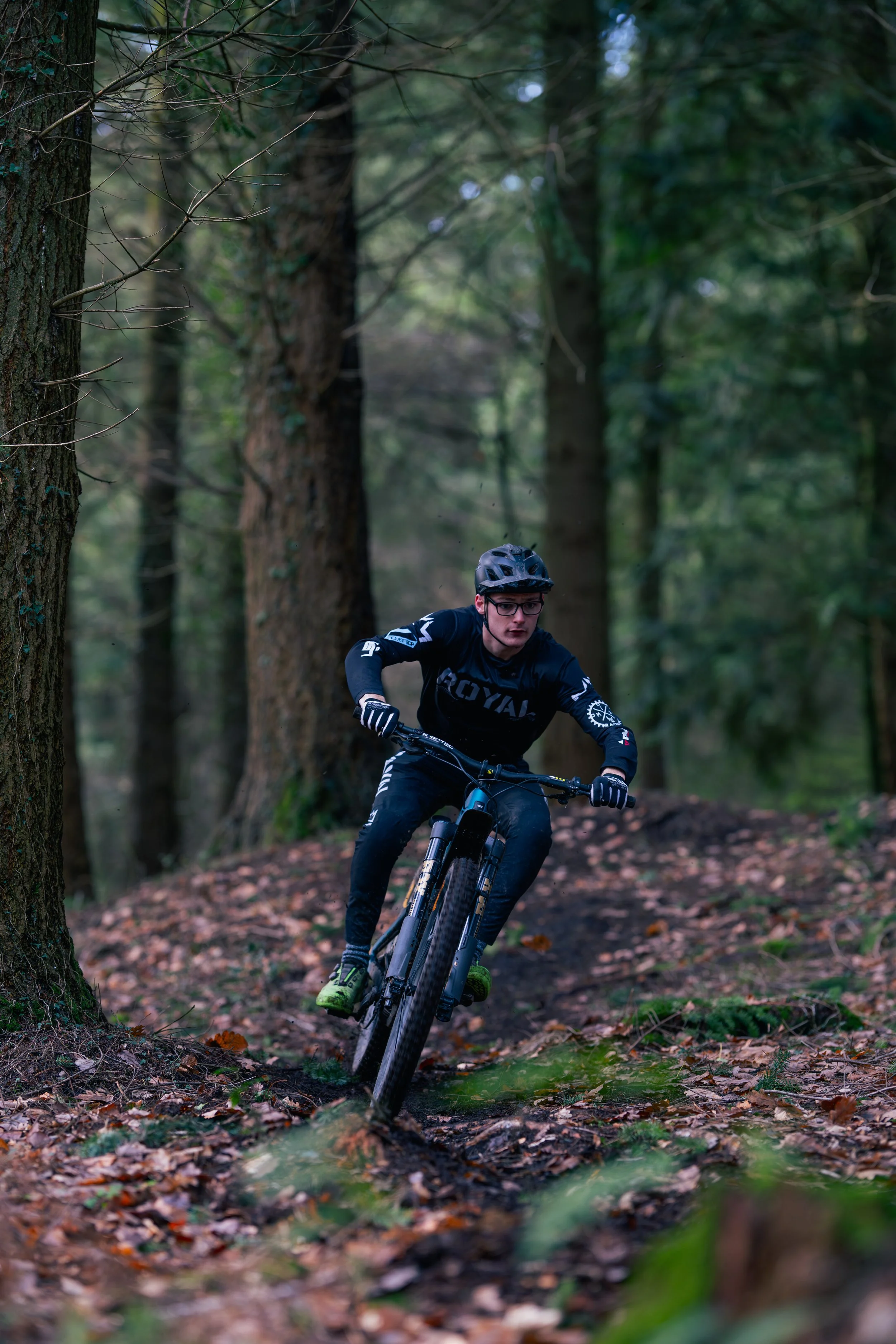 A person mountain biking on a trail in a dense forest, wearing a black helmet, glasses, and a black long-sleeve shirt.