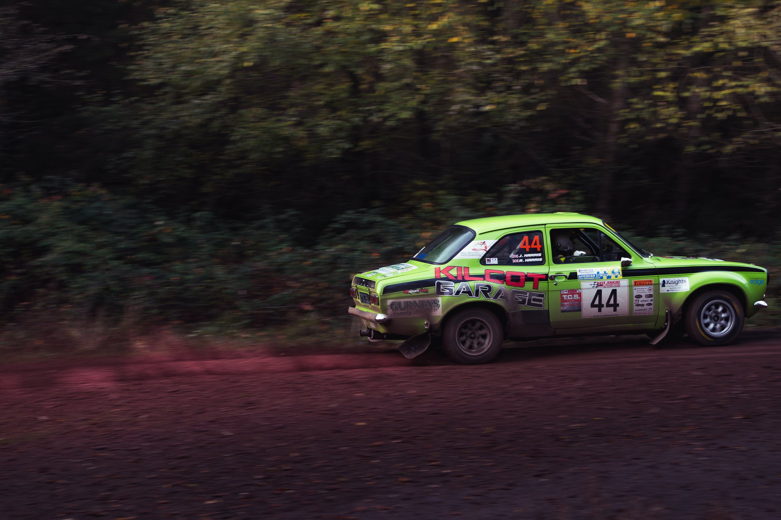 A bright green vintage rally car numbered 44 racing on a dirt track, kicking up dust with a forest background. wye dean rally 